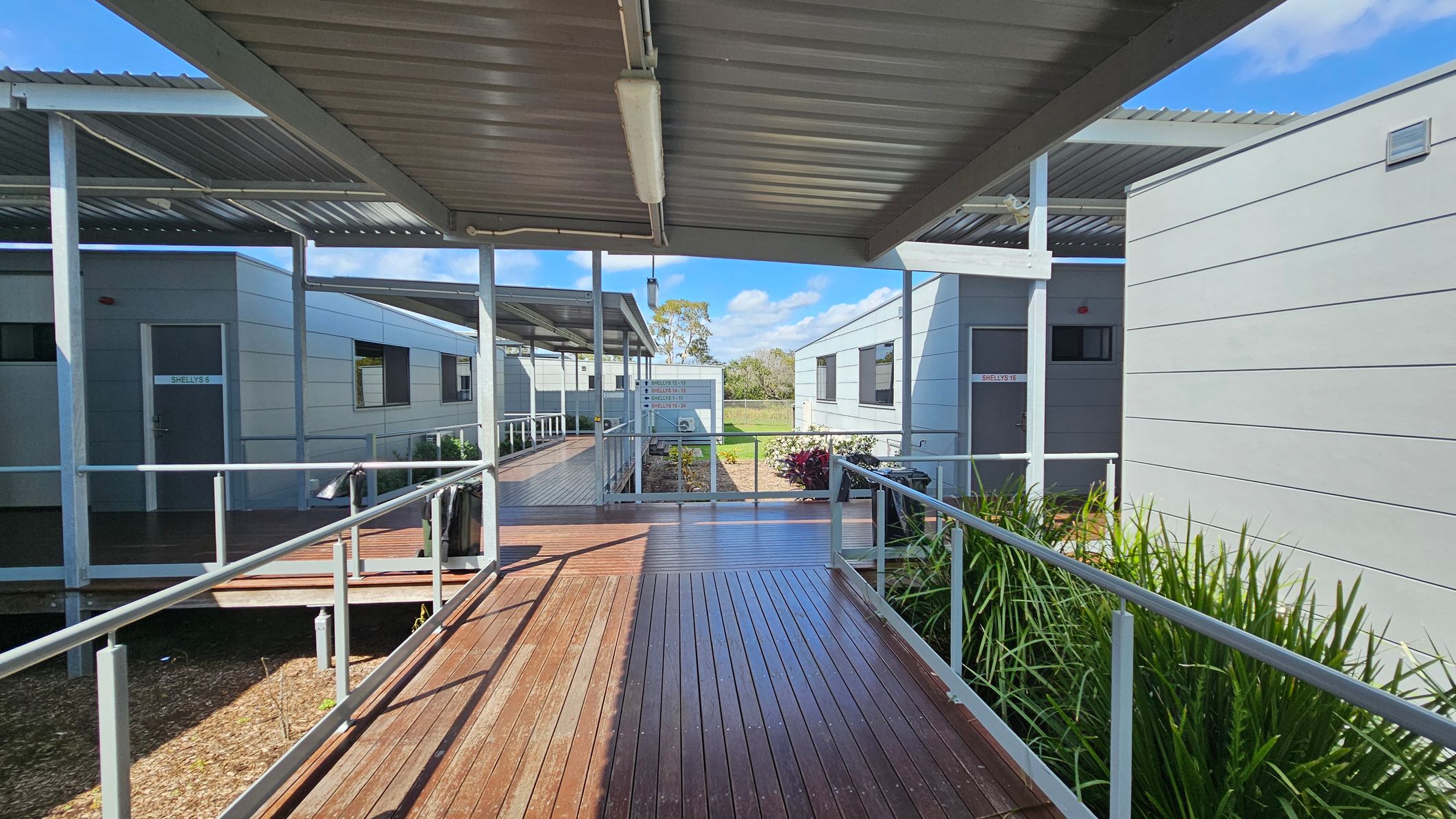Covered walkways with wooden decking connect several grey modular buildings. On the left, a door labelled "SHELLEY 5" is visible. On the right, another building has a door marked "SHELLEY 16". Metal railings line the walkways, and there are green plants along the edges. The ceiling is made of corrugated metal. In the background, a grassy area and trees are visible under a blue sky. Covered walkways with wooden decking connect several grey modular buildings. On the left, a door labelled "SHELLEY 5" is visible. On the right, another building has a door marked "SHELLEY 16". Metal railings line the walkways, and there are green plants along the edges. The ceiling is made of corrugated metal. In the background, a grassy area and trees are visible under a blue sky.