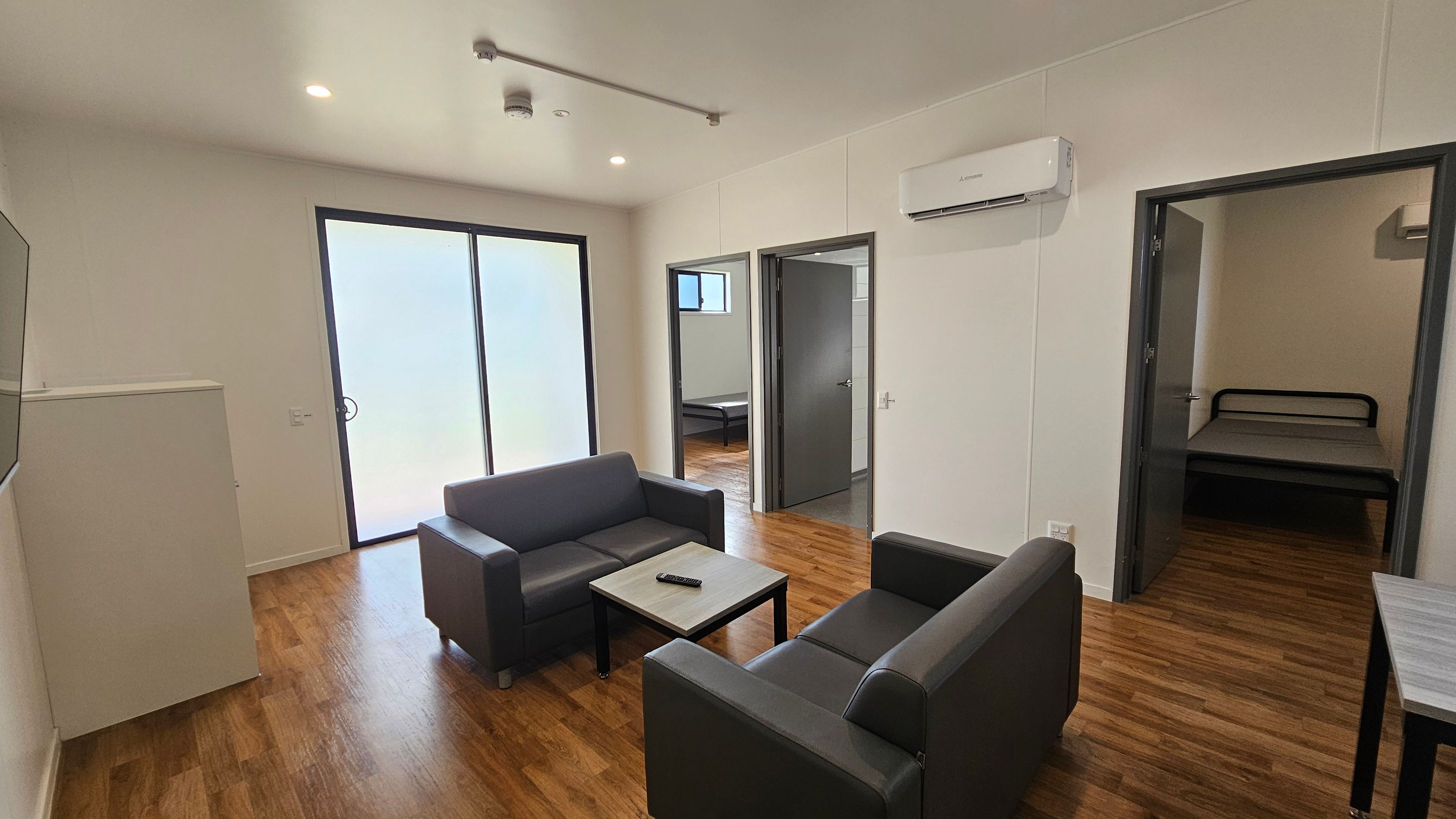 A modern living room with wooden flooring. In the centre, two dark grey sofas face each other with a small coffee table between them, holding a remote control. On the left, large sliding glass doors allow natural light in. On the right, two doorways lead to separate rooms, each containing a bed. An air conditioning unit is mounted on the wall above the right doorway. The ceiling is white with recessed lighting. A modern living room with wooden flooring. In the centre, two dark grey sofas face each other with a small coffee table between them, holding a remote control. On the left, large sliding glass doors allow natural light in. On the right, two doorways lead to separate rooms, each containing a bed. An air conditioning unit is mounted on the wall above the right doorway. The ceiling is white with recessed lighting.