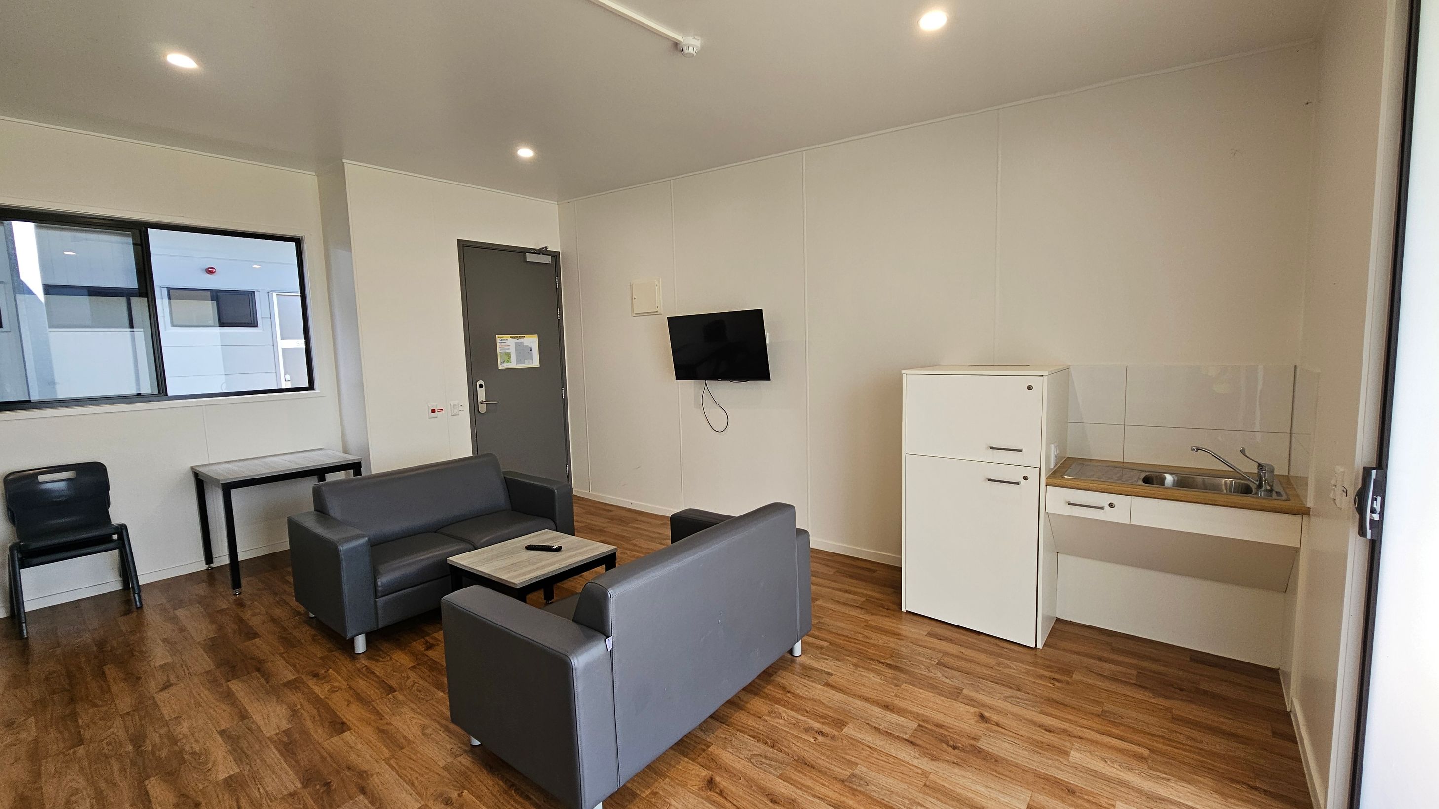 On the left, there is a dark-coloured chair next to a small table with a light wood surface. A large window with a black frame allows light into the space. To the right, a grey door leads to another area, and a television is mounted on the wall next to it. A white fridge stands beside a compact kitchen area with a sink and a countertop. The floor is covered in light wooden laminate, providing a warm tone. The ceiling is flat and painted white, with several recessed lights. On the left, there is a dark-coloured chair next to a small table with a light wood surface. A large window with a black frame allows light into the space. To the right, a grey door leads to another area, and a television is mounted on the wall next to it. A white fridge stands beside a compact kitchen area with a sink and a countertop. The floor is covered in light wooden laminate, providing a warm tone. The ceiling is flat and painted white, with several recessed lights.