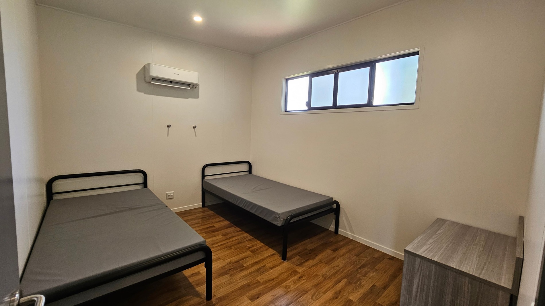On the left, a single bed with a black metal frame is positioned against the wall, covered with a grey mattress. To the right, another similar bed is set parallel, also with a grey mattress. An air conditioning unit is mounted above the beds, and two wall hooks provide additional storage. In the background, a narrow window with multiple panes allows light into the room. The floor is covered in a light wooden laminate, and the ceiling is flat with bright recessed lights. On the left, a single bed with a black metal frame is positioned against the wall, covered with a grey mattress. To the right, another similar bed is set parallel, also with a grey mattress. An air conditioning unit is mounted above the beds, and two wall hooks provide additional storage. In the background, a narrow window with multiple panes allows light into the room. The floor is covered in a light wooden laminate, and the ceiling is flat with bright recessed lights.