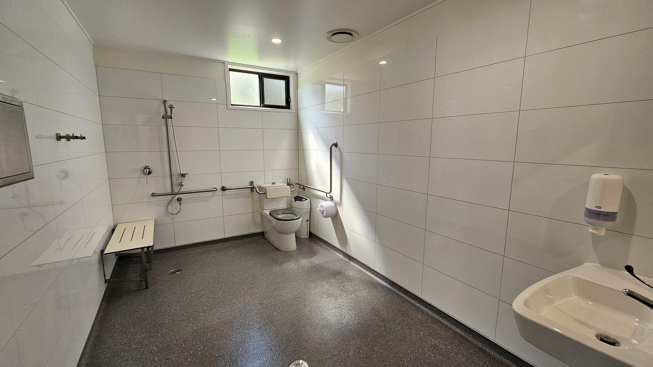 A spacious, accessible bathroom with white tiled walls and a grey speckled floor. On the left, there's a shower area with a bench and grab bars. In the centre, a toilet with additional grab bars and a small window above. On the right, a sink with a soap dispenser mounted on the wall. The ceiling is white with recessed lighting. A spacious, accessible bathroom with white tiled walls and a grey speckled floor. On the left, there's a shower area with a bench and grab bars. In the centre, a toilet with additional grab bars and a small window above. On the right, a sink with a soap dispenser mounted on the wall. The ceiling is white with recessed lighting.