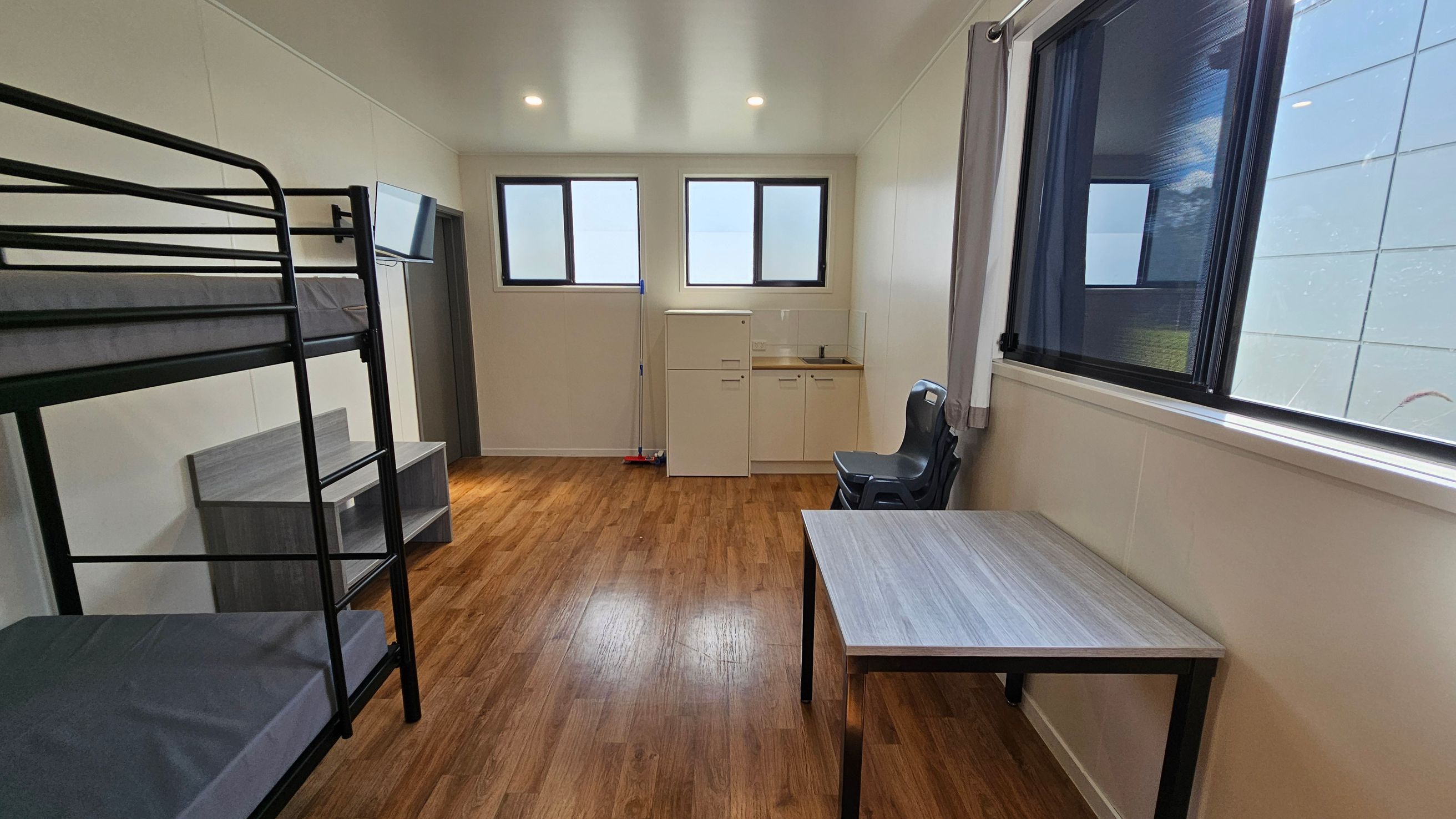 On the left, a black metal bunk bed occupies the corner, with a small grey bedside table beside it. To the right, a grey plastic chair sits adjacent to a light wood table. A compact kitchenette is positioned against the wall, featuring a sink and storage cabinet below. In the background, two rectangular windows allow natural light into the room, partially covered by light curtains. The floor is finished in a light wooden laminate, and the ceiling is flat and white, fitted with recessed light. On the left, a black metal bunk bed occupies the corner, with a small grey bedside table beside it. To the right, a grey plastic chair sits adjacent to a light wood table. A compact kitchenette is positioned against the wall, featuring a sink and storage cabinet below. In the background, two rectangular windows allow natural light into the room, partially covered by light curtains. The floor is finished in a light wooden laminate, and the ceiling is flat and white, fitted with recessed light.