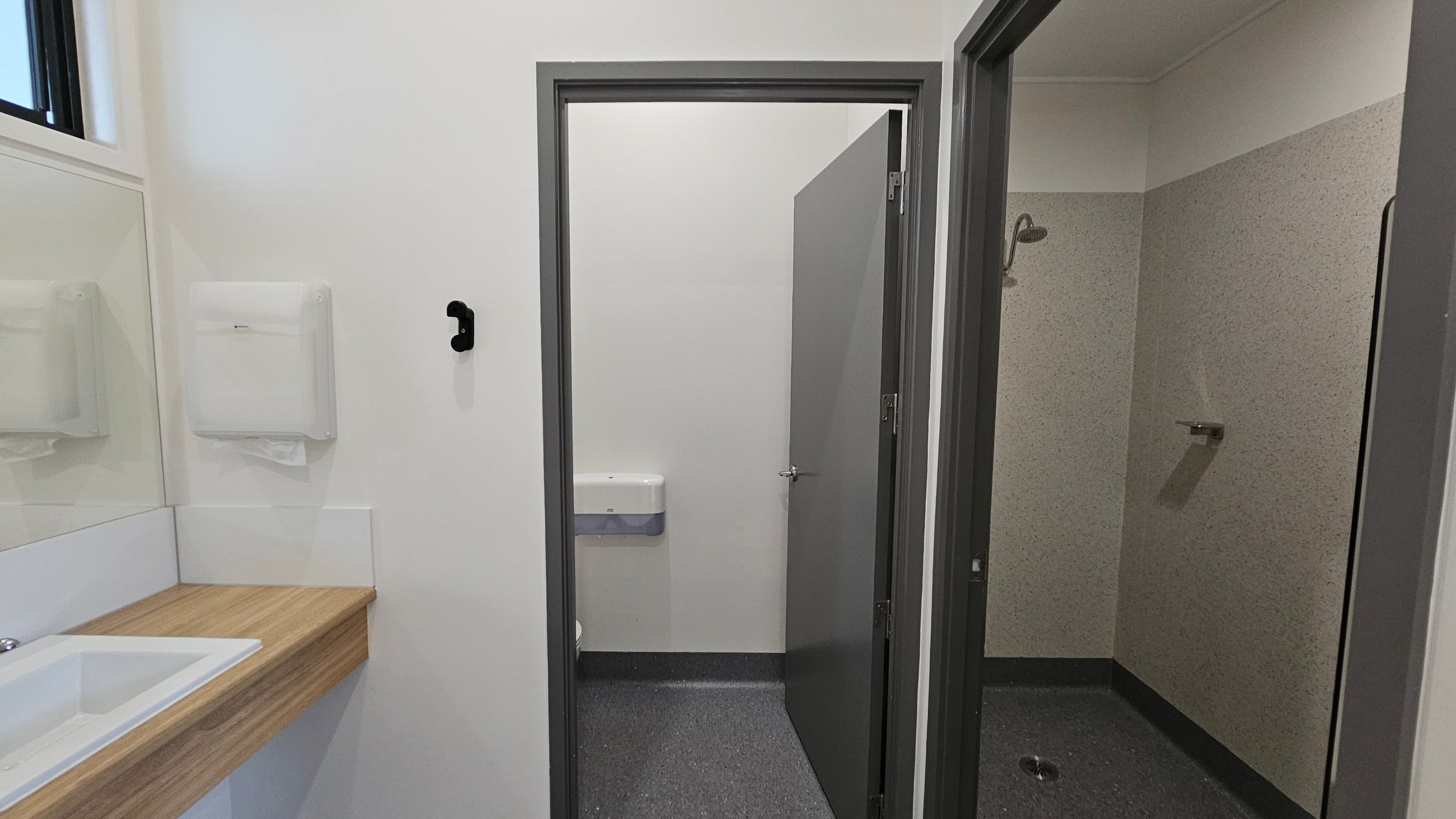 To the left, a wooden countertop with a white sink is located beneath a large mirror, with a paper towel dispenser mounted on the wall above it. There is a black hook on the wall nearby. On the right, an open grey door leads to a shower area, which features a wall-mounted showerhead and a small drain on the floor. A second door, also grey, is slightly ajar, revealing a toilet with a flush mechanism visible. The floor is a dark grey, textured surface, and the ceiling is flat and white. To the left, a wooden countertop with a white sink is located beneath a large mirror, with a paper towel dispenser mounted on the wall above it. There is a black hook on the wall nearby. On the right, an open grey door leads to a shower area, which features a wall-mounted showerhead and a small drain on the floor. A second door, also grey, is slightly ajar, revealing a toilet with a flush mechanism visible. The floor is a dark grey, textured surface, and the ceiling is flat and white.
