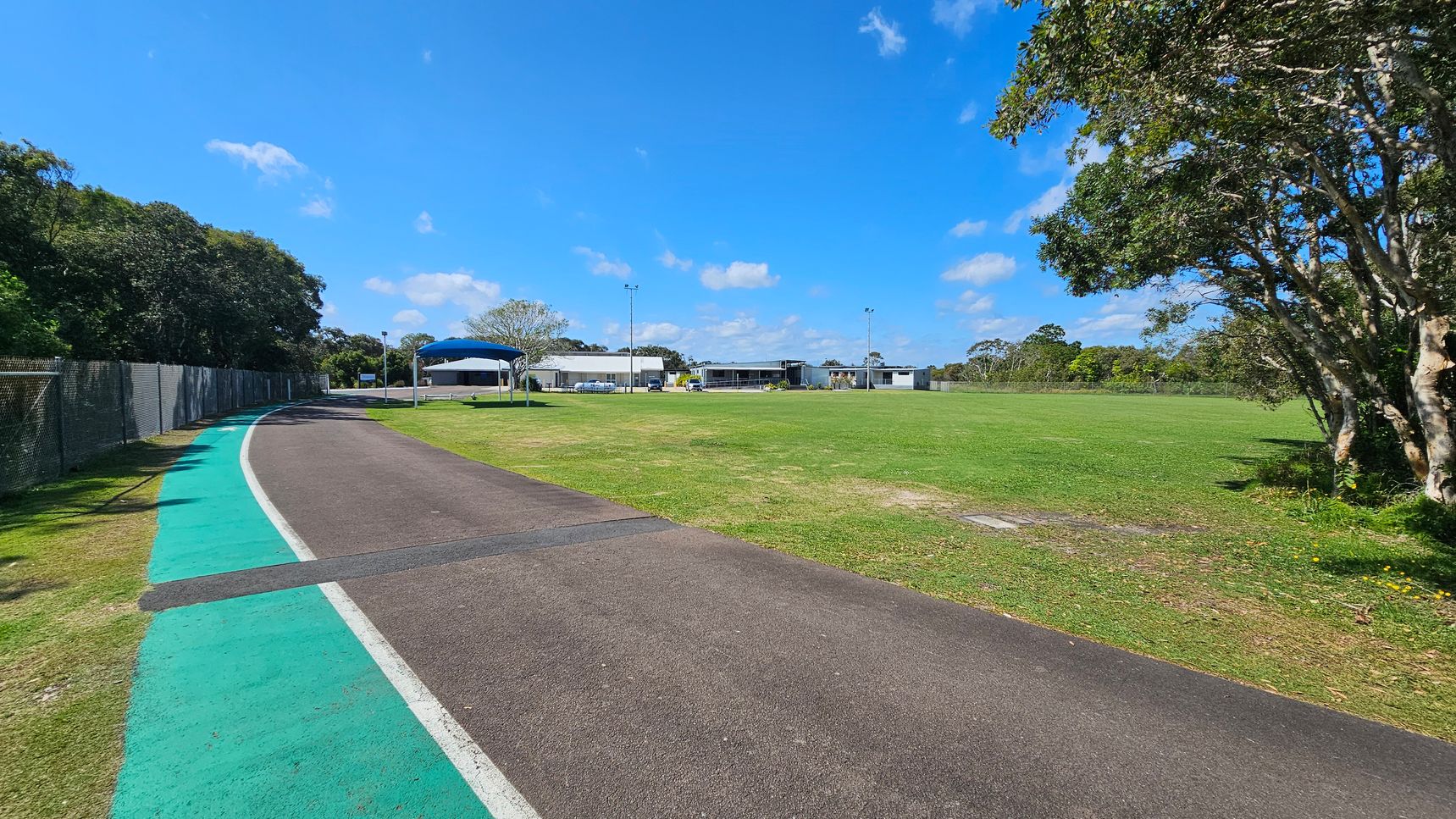 On the left, a narrow path curves alongside a wire fence, with a green strip adjacent to the asphalt surface. The path leads towards a series of buildings in the background, including one with a blue roof. To the right, there is a wide expanse of well-groomed grass. The ground is primarily dark asphalt with a light-coloured border, transitioning to a smooth grassy area. On the left, a narrow path curves alongside a wire fence, with a green strip adjacent to the asphalt surface. The path leads towards a series of buildings in the background, including one with a blue roof. To the right, there is a wide expanse of well-groomed grass. The ground is primarily dark asphalt with a light-coloured border, transitioning to a smooth grassy area.