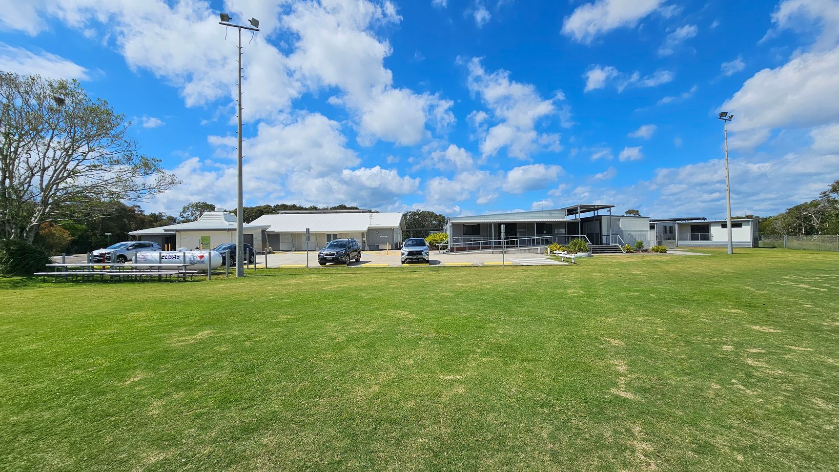To the left, there are several parked cars near a small structure, with benches lined up parallel to the grass. On the right, a larger building features a raised deck area with steps leading up to it. The floor is a manicured expanse of green grass extending across the foreground, with a light-coloured concrete path visible near the buildings. In the background, there are utility poles and trees providing some greenery around the area. e. To the left, there are several parked cars near a small structure, with benches lined up parallel to the grass. On the right, a larger building features a raised deck area with steps leading up to it. The floor is a manicured expanse of green grass extending across the foreground, with a light-coloured concrete path visible near the buildings. In the background, there are utility poles and trees providing some greenery around the area. e.