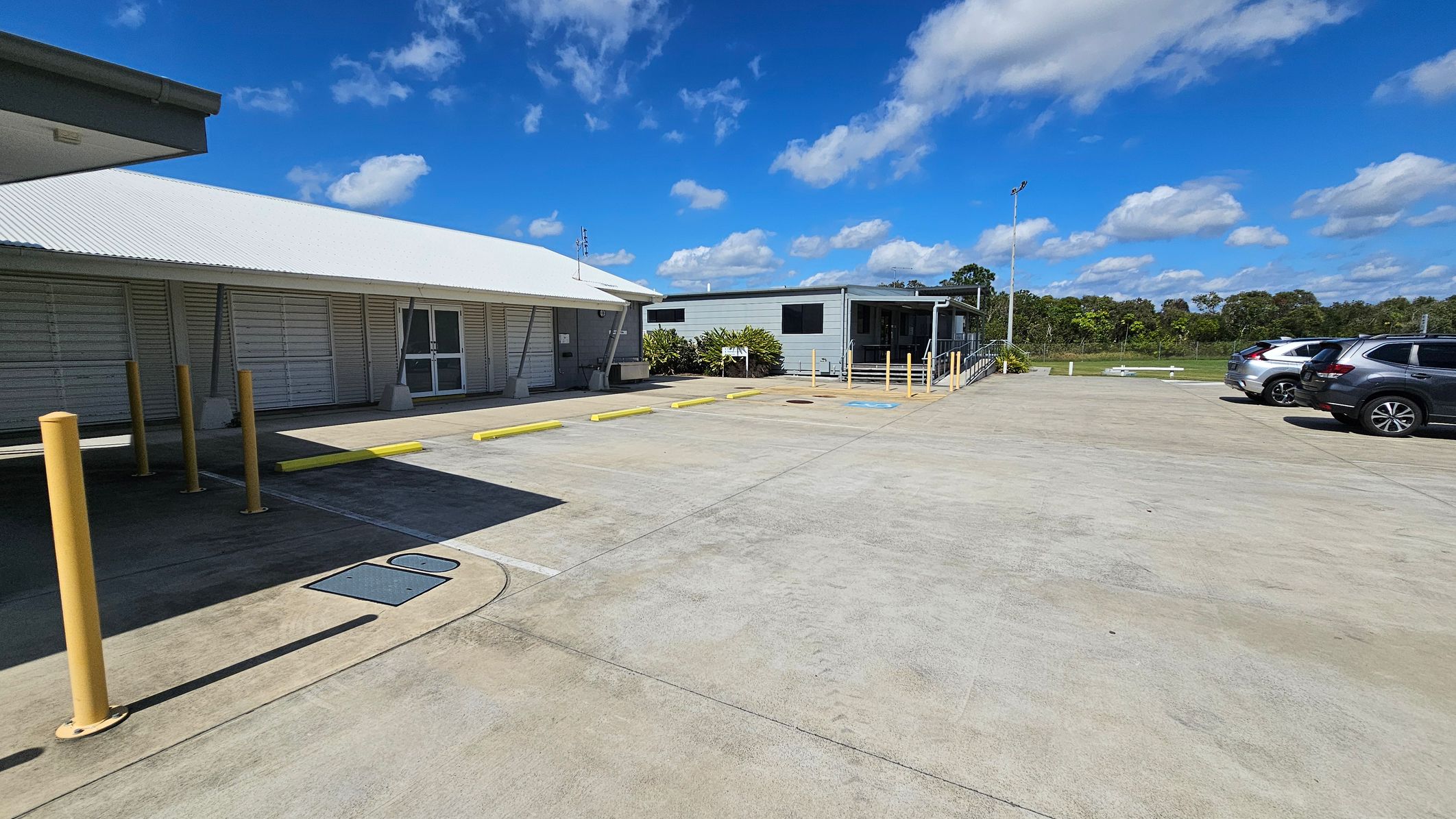 On the left, there is a building with a light-coloured wooden façade and a sloping roof, featuring a set of double doors. In front, yellow bollards line the edge of a concrete parking area. To the right, another structure with a flat roof has a small set of stairs leading up to it, surrounded by some greenery. A few parked cars are visible, occupying a smooth, light grey concrete surface. On the left, there is a building with a light-coloured wooden façade and a sloping roof, featuring a set of double doors. In front, yellow bollards line the edge of a concrete parking area. To the right, another structure with a flat roof has a small set of stairs leading up to it, surrounded by some greenery. A few parked cars are visible, occupying a smooth, light grey concrete surface.