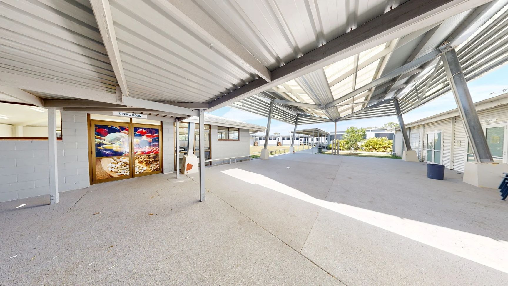 On the left, there is a door marked "DINING ROOM" with a large decorative glass panel featuring colourful fish. Adjacent to it, a grey wall with a window is visible. To the right, additional buildings extend into the background, showing more windows and entrances. The floor is a smooth, pale concrete surface, reflecting light. The ceiling overhead is made of corrugated metal, supported by steel beams, creating a spacious covered area. On the left, there is a door marked "DINING ROOM" with a large decorative glass panel featuring colourful fish. Adjacent to it, a grey wall with a window is visible. To the right, additional buildings extend into the background, showing more windows and entrances. The floor is a smooth, pale concrete surface, reflecting light. The ceiling overhead is made of corrugated metal, supported by steel beams, creating a spacious covered area.