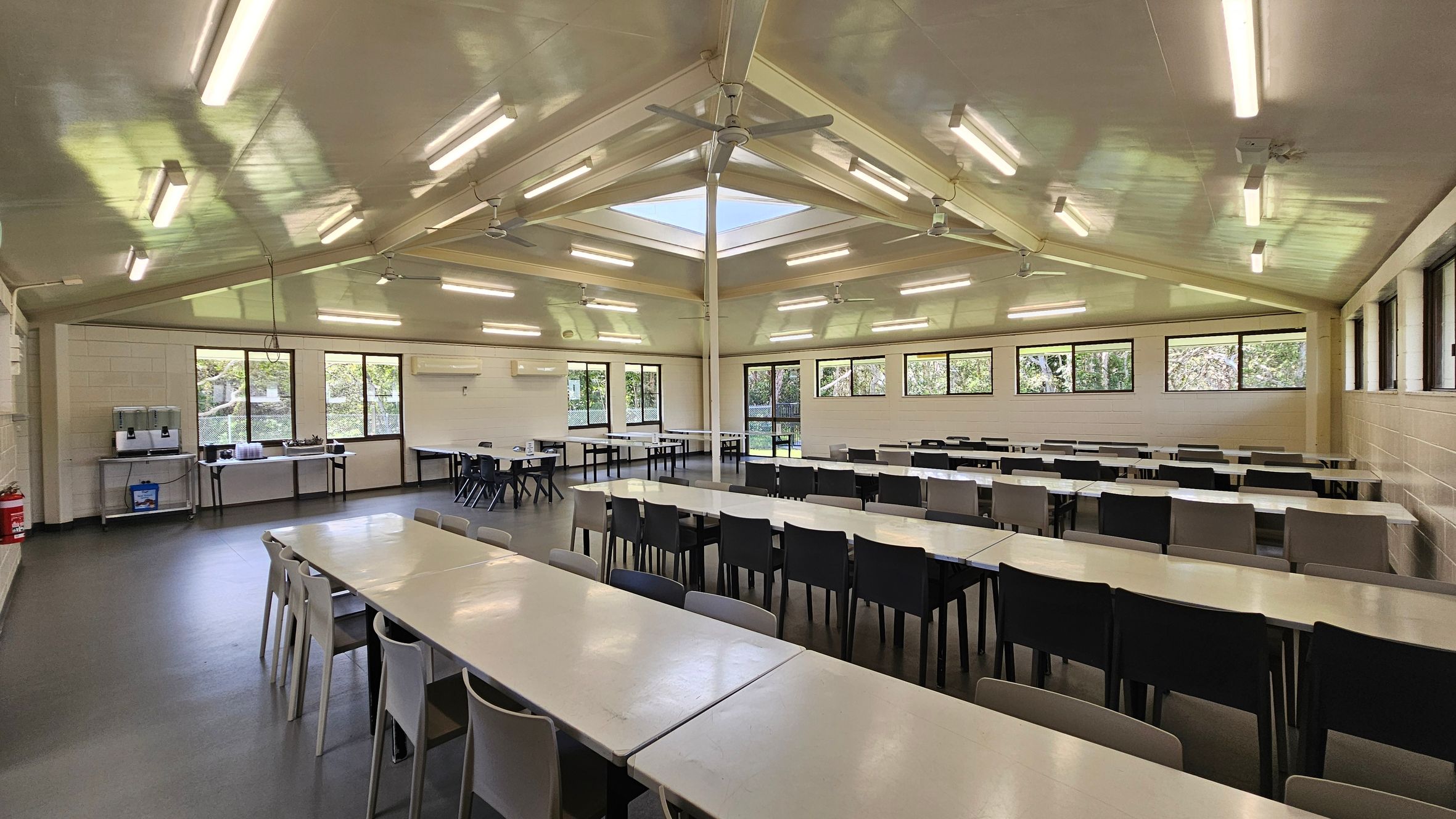 To the left, there are two large windows allowing natural light to enter, with a serving area featuring a water dispenser and trays. Adjacent is a wall-mounted air conditioning unit. On the right, additional windows extend along the wall, providing views of greenery outside. In the middle, multiple long tables with white surfaces are arranged, surrounded by black and grey chairs. The floor is a smooth dark surface, and the ceiling is flat with ceiling fans and fluorescent lights. To the left, there are two large windows allowing natural light to enter, with a serving area featuring a water dispenser and trays. Adjacent is a wall-mounted air conditioning unit. On the right, additional windows extend along the wall, providing views of greenery outside. In the middle, multiple long tables with white surfaces are arranged, surrounded by black and grey chairs. The floor is a smooth dark surface, and the ceiling is flat with ceiling fans and fluorescent lights.