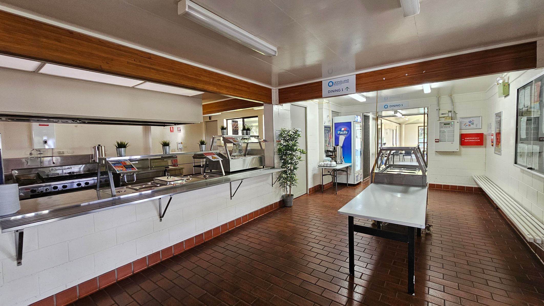 On the left, there is a serving counter with metal fixtures and several trays arranged neatly, alongside potted plants for decoration. A small window reveals more kitchen space behind the counter. To the right, a vending machine and a table with hot drink dispensers are positioned against the wall. In the middle, a stainless steel table stands ready for food preparation. The floor features dark brown tiles arranged in a checkerboard pattern, while the ceiling is flat with fluorescent lights. On the left, there is a serving counter with metal fixtures and several trays arranged neatly, alongside potted plants for decoration. A small window reveals more kitchen space behind the counter. To the right, a vending machine and a table with hot drink dispensers are positioned against the wall. In the middle, a stainless steel table stands ready for food preparation. The floor features dark brown tiles arranged in a checkerboard pattern, while the ceiling is flat with fluorescent lights.