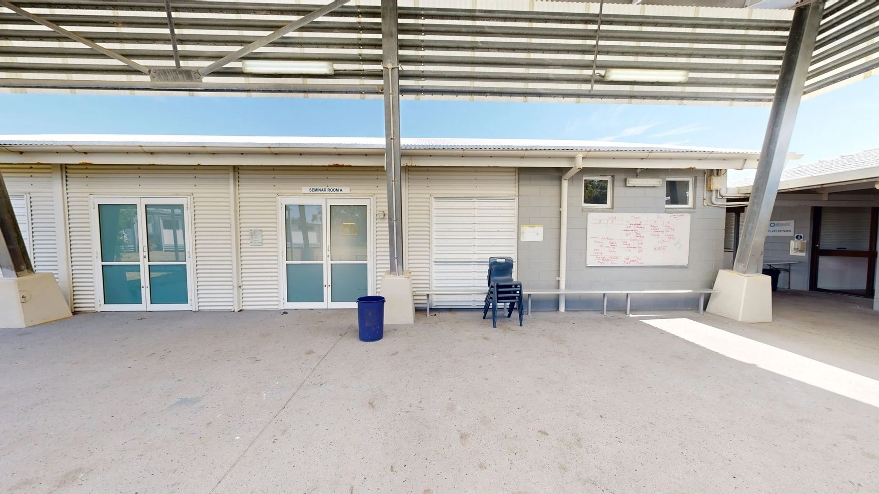 On the left, two glass doors marked "Seminar Room A" lead into a building with a light-coloured, corrugated exterior. Next to it, a wall with a whiteboard covered in notes is visible. To the right, a second entrance leads into a different room, with a small window above it. A blue bin stands on the concrete floor, alongside a stack of chairs. The ground is a smooth, pale concrete surface. Above, a metal roof supported by beams provides shade, with fluorescent lights mounted underneath. On the left, two glass doors marked "Seminar Room A" lead into a building with a light-coloured, corrugated exterior. Next to it, a wall with a whiteboard covered in notes is visible. To the right, a second entrance leads into a different room, with a small window above it. A blue bin stands on the concrete floor, alongside a stack of chairs. The ground is a smooth, pale concrete surface. Above, a metal roof supported by beams provides shade, with fluorescent lights mounted underneath.