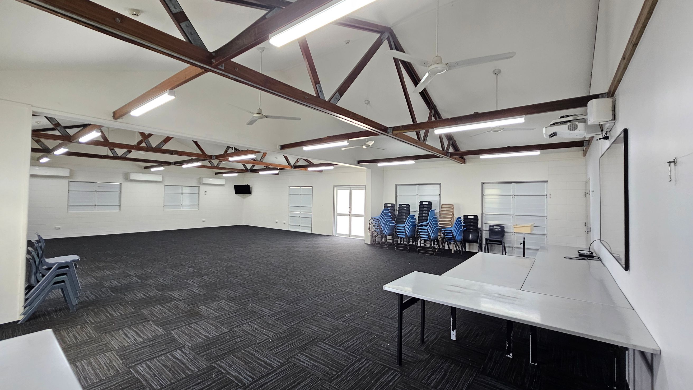 To the left, a stack of grey chairs is arranged against a white wall, beside a doorway. On the right, a row of windows lets in light, with a wall-mounted television just above. A large whiteboard is mounted nearby. In the middle, a rectangular table with a smooth surface is positioned at an angle, facing the room. The floor features dark carpet tiles arranged in a patterned layout. The ceiling has exposed wooden beams, with ceiling fans and fluorescent lights evenly spaced throughout. To the left, a stack of grey chairs is arranged against a white wall, beside a doorway. On the right, a row of windows lets in light, with a wall-mounted television just above. A large whiteboard is mounted nearby. In the middle, a rectangular table with a smooth surface is positioned at an angle, facing the room. The floor features dark carpet tiles arranged in a patterned layout. The ceiling has exposed wooden beams, with ceiling fans and fluorescent lights evenly spaced throughout.