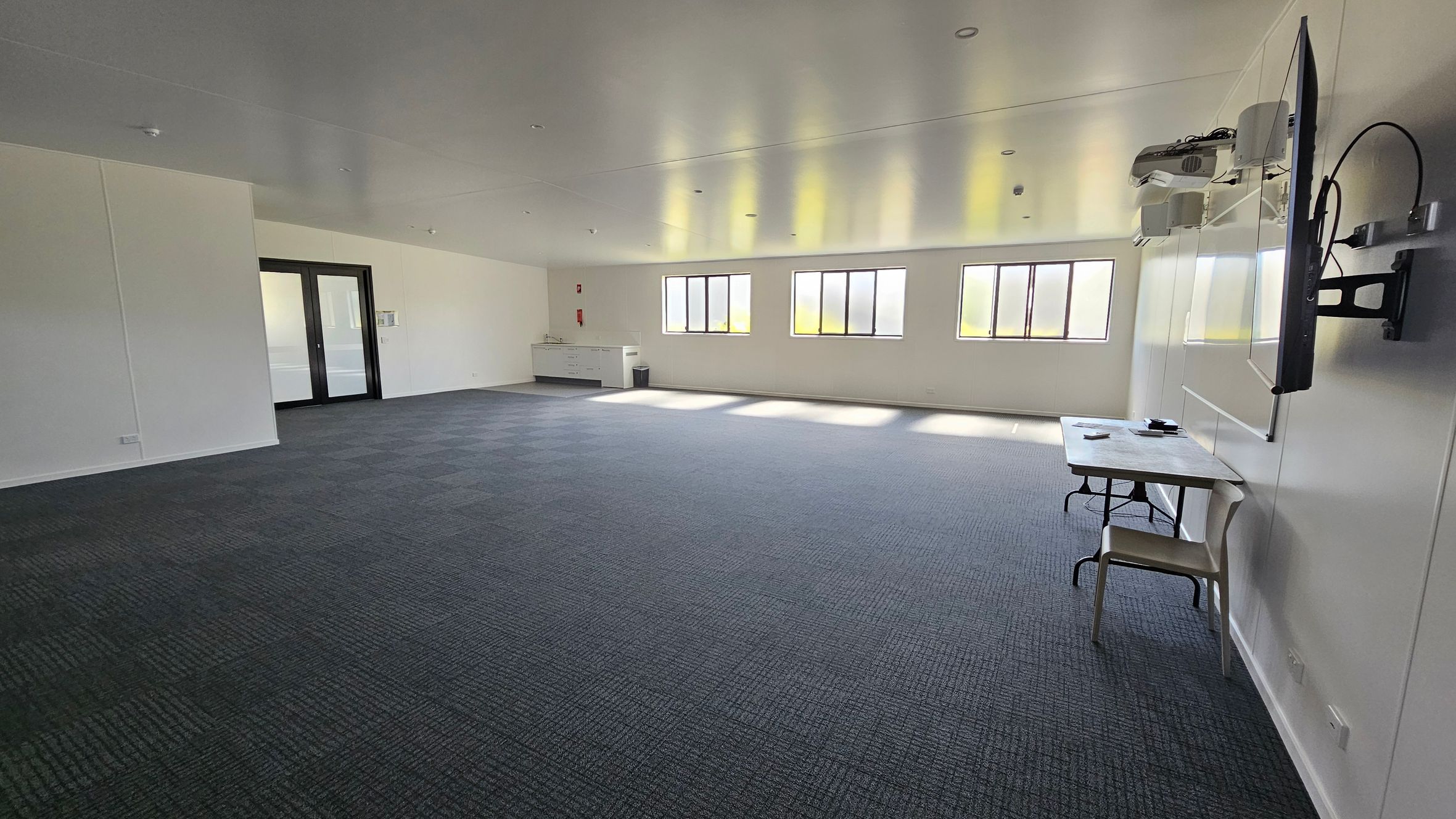 On the left, large double doors lead to another space, with a wall-mounted notice board beside them. A service counter is situated nearby, featuring a sink and storage below. To the right, several tall windows provide natural light, with a flat-screen television mounted on the wall. In the middle, a simple table is set against the wall with a chair beside it. The floor is covered in dark blue carpet with a textured pattern. The ceiling is flat and smooth, fitted with recessed lighting. On the left, large double doors lead to another space, with a wall-mounted notice board beside them. A service counter is situated nearby, featuring a sink and storage below. To the right, several tall windows provide natural light, with a flat-screen television mounted on the wall. In the middle, a simple table is set against the wall with a chair beside it. The floor is covered in dark blue carpet with a textured pattern. The ceiling is flat and smooth, fitted with recessed lighting.