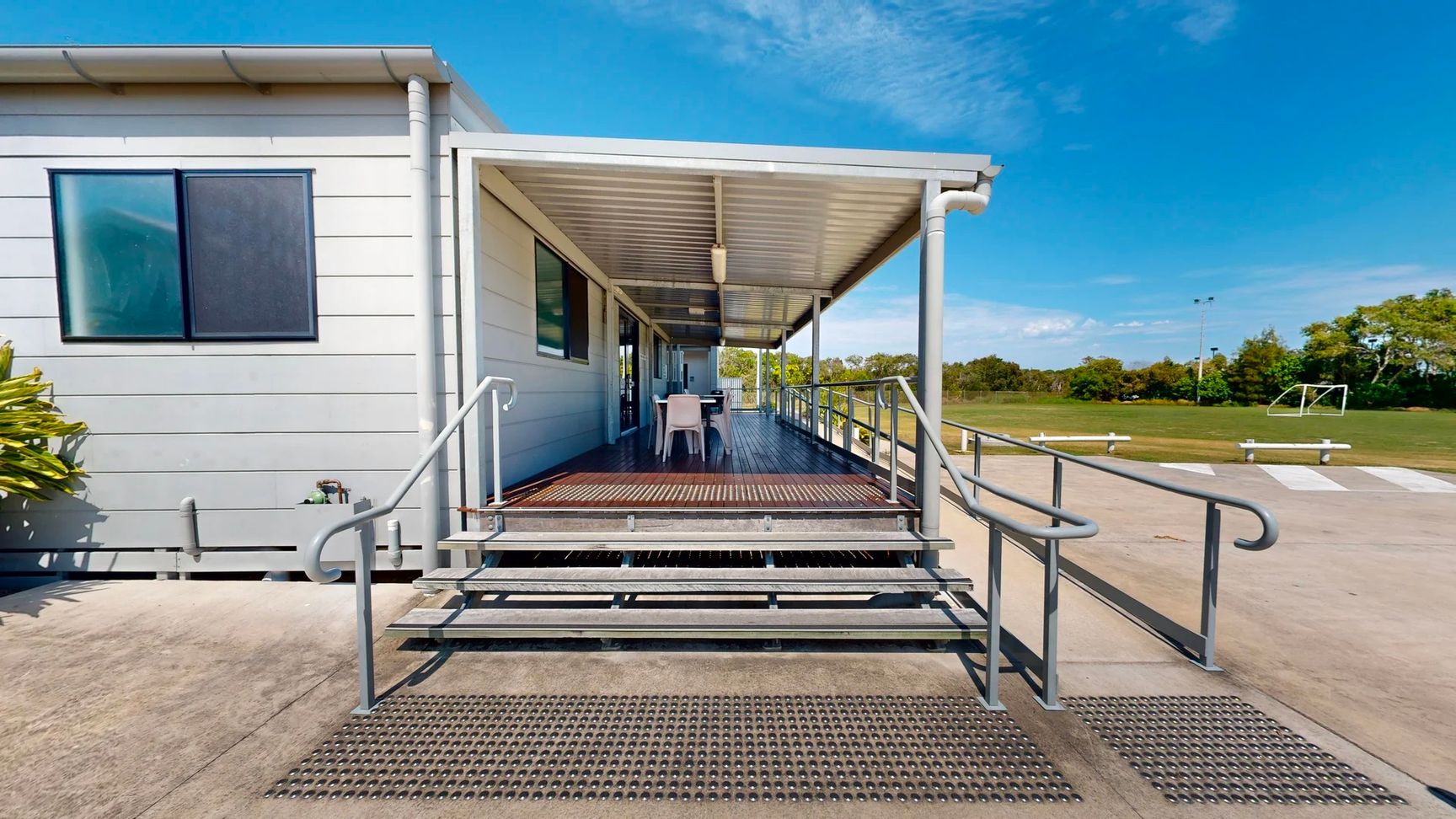 On the left, a small window with a dark frame is visible, alongside a light-coloured wall of a building. A cluster of green plants is situated next to the steps. To the right, a set of stainless steel rails leads to a deck area with a table and chairs under a corrugated metal roof. The foreground features a wide, grey concrete pathway with a textured surface, while the background reveals a grassy area with white benches and a goalpost in the distance. On the left, a small window with a dark frame is visible, alongside a light-coloured wall of a building. A cluster of green plants is situated next to the steps. To the right, a set of stainless steel rails leads to a deck area with a table and chairs under a corrugated metal roof. The foreground features a wide, grey concrete pathway with a textured surface, while the background reveals a grassy area with white benches and a goalpost in the distance.