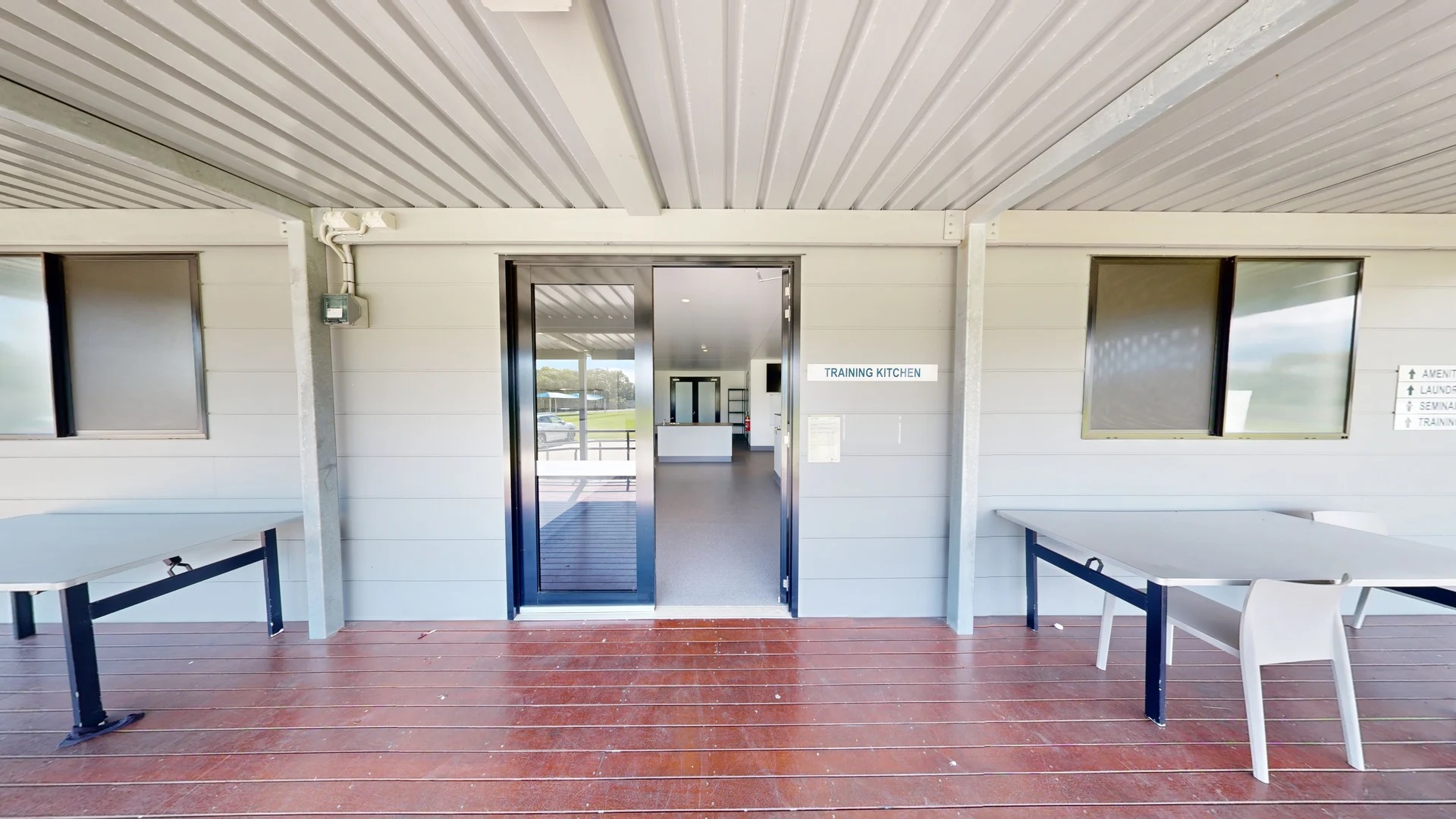 To the left, a grey table with a black frame stands, partially shaded by the overhang. Adjacent to it, a window allows light into the interior space. On the right, another grey table is positioned similarly, with a white chair beside it. In the middle, a set of sliding glass doors leads into a room labelled "TRAINING KITCHEN." The floor consists of polished wooden decking, and the ceiling is flat with a corrugated metal design, featuring built-in lights that illuminate the entryway. To the left, a grey table with a black frame stands, partially shaded by the overhang. Adjacent to it, a window allows light into the interior space. On the right, another grey table is positioned similarly, with a white chair beside it. In the middle, a set of sliding glass doors leads into a room labelled "TRAINING KITCHEN." The floor consists of polished wooden decking, and the ceiling is flat with a corrugated metal design, featuring built-in lights that illuminate the entryway.