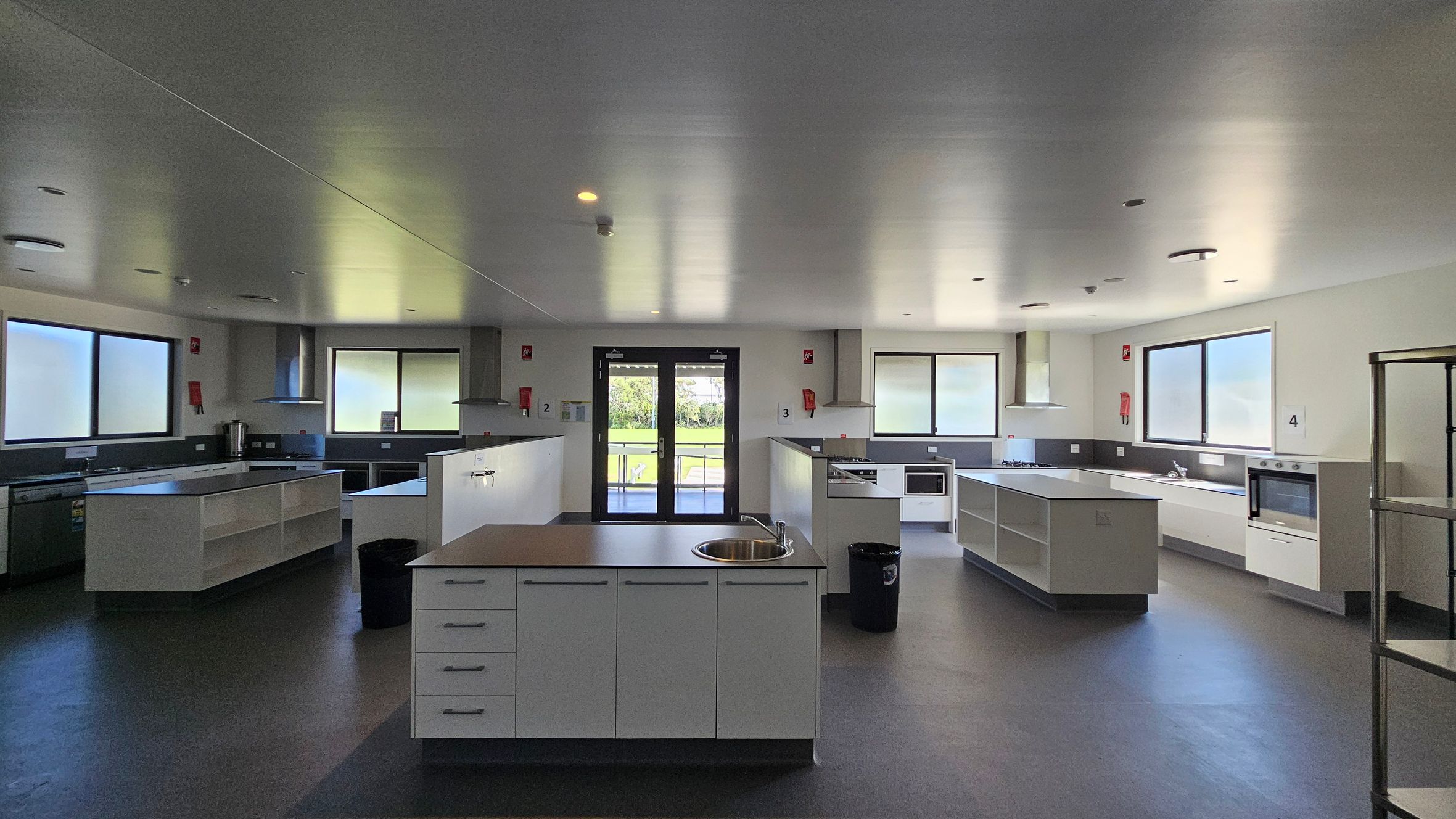 On the left, a stainless steel kitchen unit includes a sink and countertops, with cabinets above for storage. To the right, another cooking station features a hob and an oven, alongside more countertop space. In the middle, an island with a circular sink is surrounded by drawers. The floor is a dark grey, smooth vinyl, while the ceiling is flat and painted white, with recessed lights evenly distributed. Large windows allow light into the room, and a set of double doors leads outside. On the left, a stainless steel kitchen unit includes a sink and countertops, with cabinets above for storage. To the right, another cooking station features a hob and an oven, alongside more countertop space. In the middle, an island with a circular sink is surrounded by drawers. The floor is a dark grey, smooth vinyl, while the ceiling is flat and painted white, with recessed lights evenly distributed. Large windows allow light into the room, and a set of double doors leads outside.