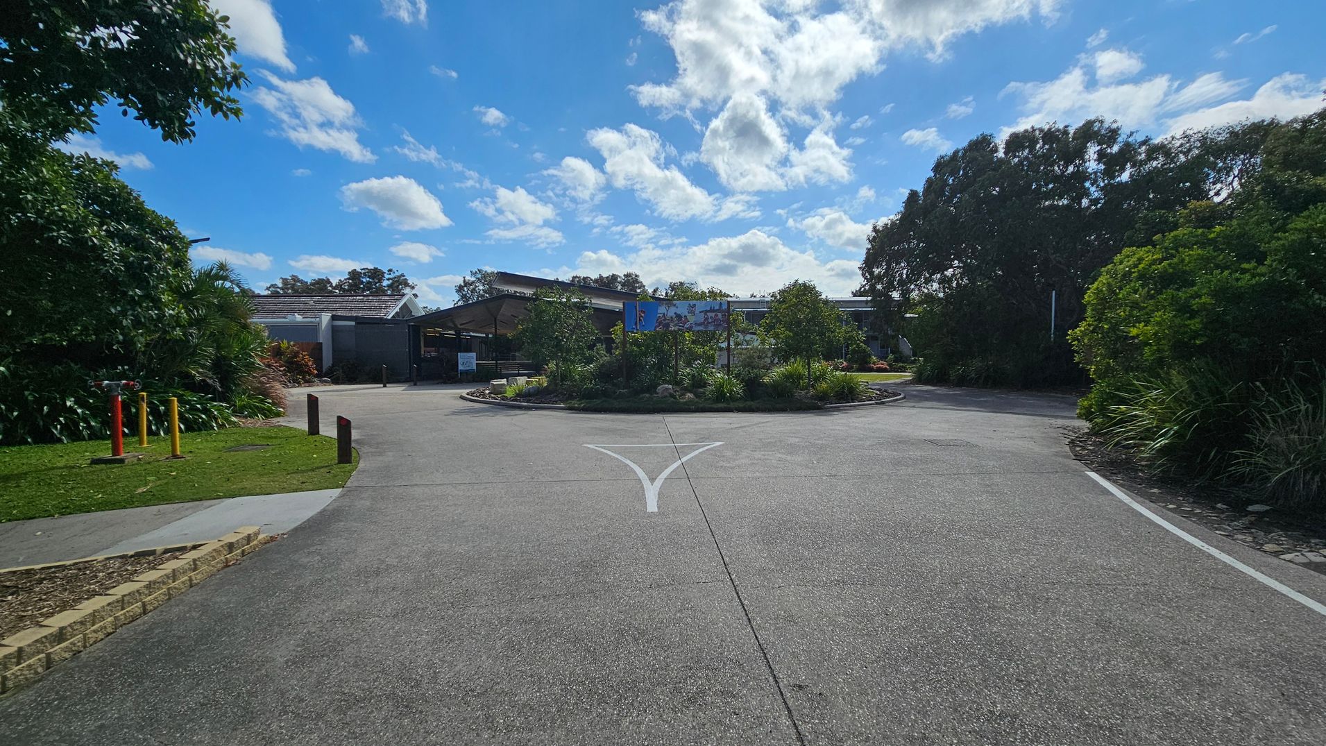 A paved road leads to a roundabout with lush greenery and small trees in the centre. On the left, there are colourful bollards and a grassy area with dense foliage. On the right, more trees and bushes line the road. In the background, a building with a sloped roof and a large sign is visible, partially obscured by the vegetation. The sky is filled with scattered clouds. A paved road leads to a roundabout with lush greenery and small trees in the centre. On the left, there are colourful bollards and a grassy area with dense foliage. On the right, more trees and bushes line the road. In the background, a building with a sloped roof and a large sign is visible, partially obscured by the vegetation. The sky is filled with scattered clouds.