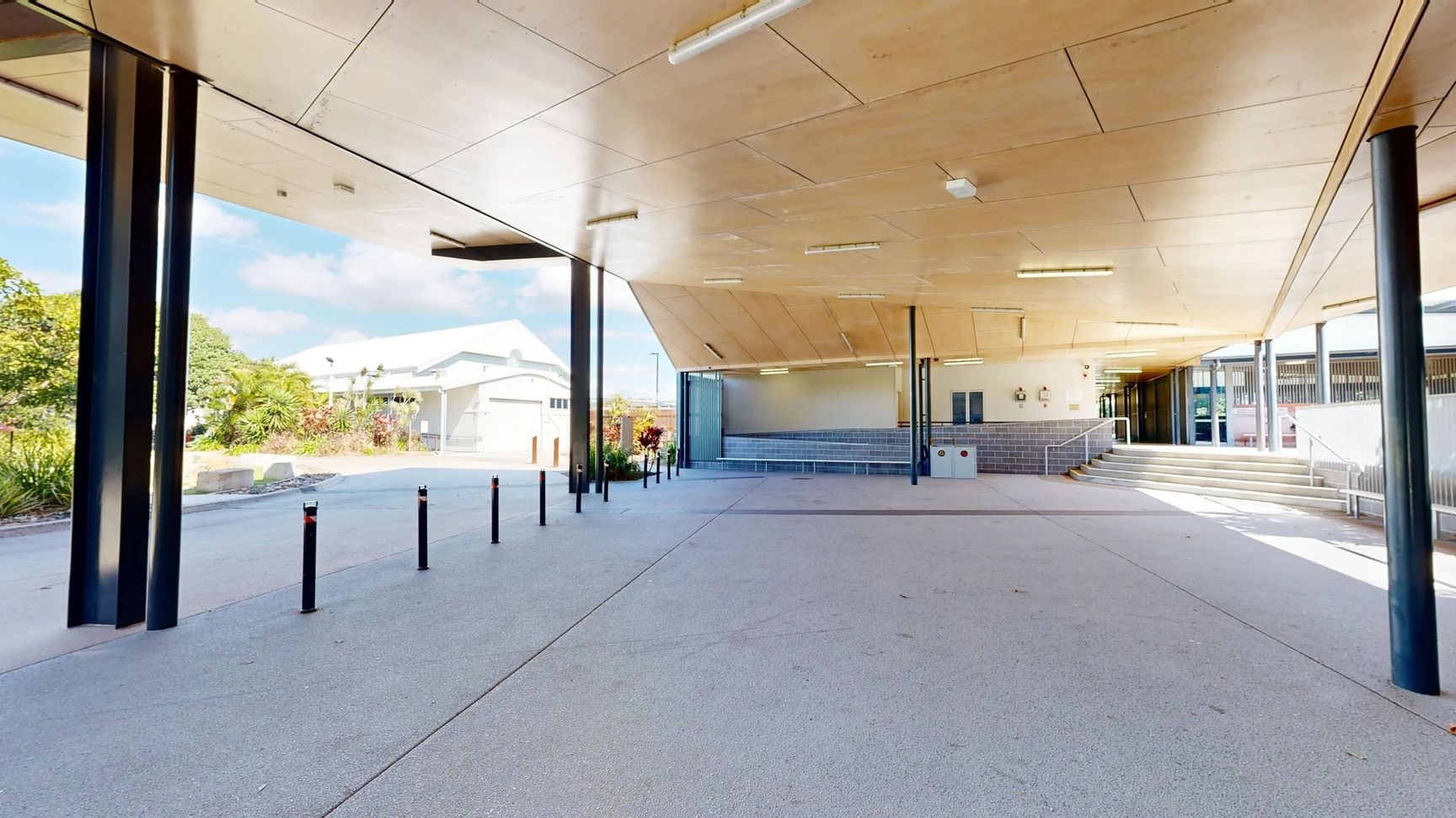 The area features a smooth concrete floor, extending underneath a canopy with a wooden panel ceiling supported by black metal columns. On the left, lush greenery and shrubs are visible beyond a row of low black bollards. In the background, a building with a peaked roof is partly visible. To the right, a set of steps leads up to a higher level, with a low wall behind it. There are several benches against the wall, and wall-mounted features can be seen as well. The area features a smooth concrete floor, extending underneath a canopy with a wooden panel ceiling supported by black metal columns. On the left, lush greenery and shrubs are visible beyond a row of low black bollards. In the background, a building with a peaked roof is partly visible. To the right, a set of steps leads up to a higher level, with a low wall behind it. There are several benches against the wall, and wall-mounted features can be seen as well.