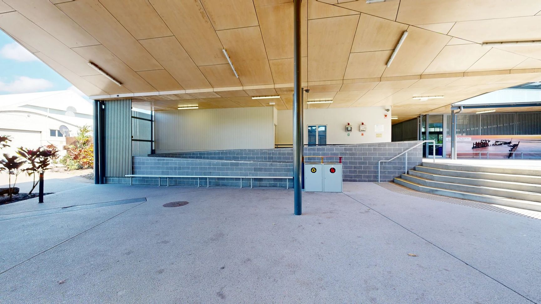 The area features a smooth, light grey concrete floor extending across the open space. On the left, a row of low shrubs adds greenery beside a vertical panel of grey cladding, while on the right, there are several steps leading up to a raised platform. The back wall has a panel with windows and two fire safety equipment units mounted near a white wall. The ceiling above is constructed of wooden panels, fitted with overhead lights , supported by tall, black metal columns in the foreground. The area features a smooth, light grey concrete floor extending across the open space. On the left, a row of low shrubs adds greenery beside a vertical panel of grey cladding, while on the right, there are several steps leading up to a raised platform. The back wall has a panel with windows and two fire safety equipment units mounted near a white wall. The ceiling above is constructed of wooden panels, fitted with overhead lights , supported by tall, black metal columns in the foreground.
