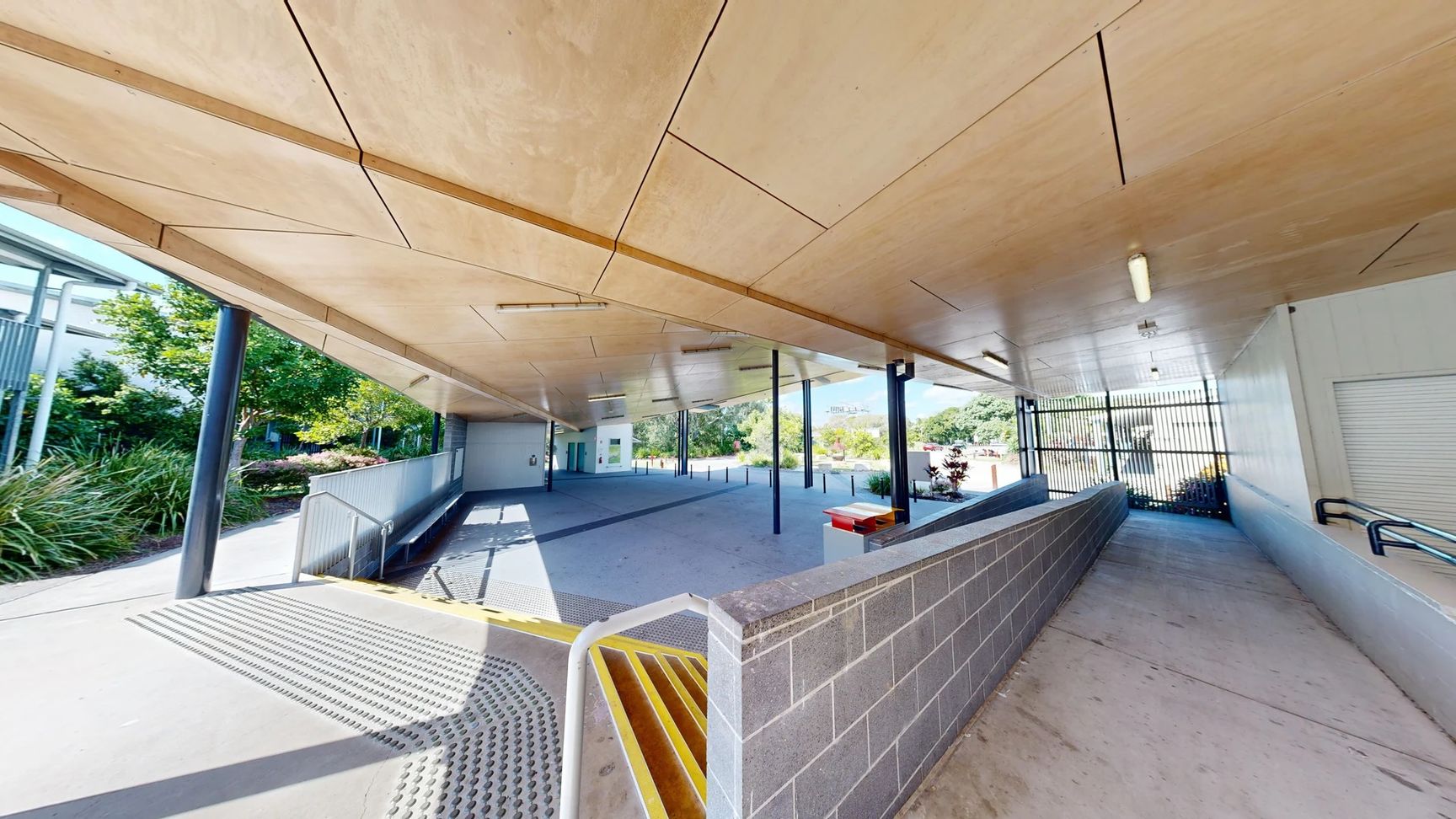 The area features a smooth concrete floor with a slight gradient leading towards a loading ramp on the left side, where a textured yellow edge marks the transition. On the right, a wall of grey bricks rises beside a metal railing, leading to a more enclosed space. The ceiling is wooden, with panels and overhead lights positioned evenly along its surface, supported by black metallic columns. In the background, greenery is visible through glass doors. The area features a smooth concrete floor with a slight gradient leading towards a loading ramp on the left side, where a textured yellow edge marks the transition. On the right, a wall of grey bricks rises beside a metal railing, leading to a more enclosed space. The ceiling is wooden, with panels and overhead lights positioned evenly along its surface, supported by black metallic columns. In the background, greenery is visible through glass doors.