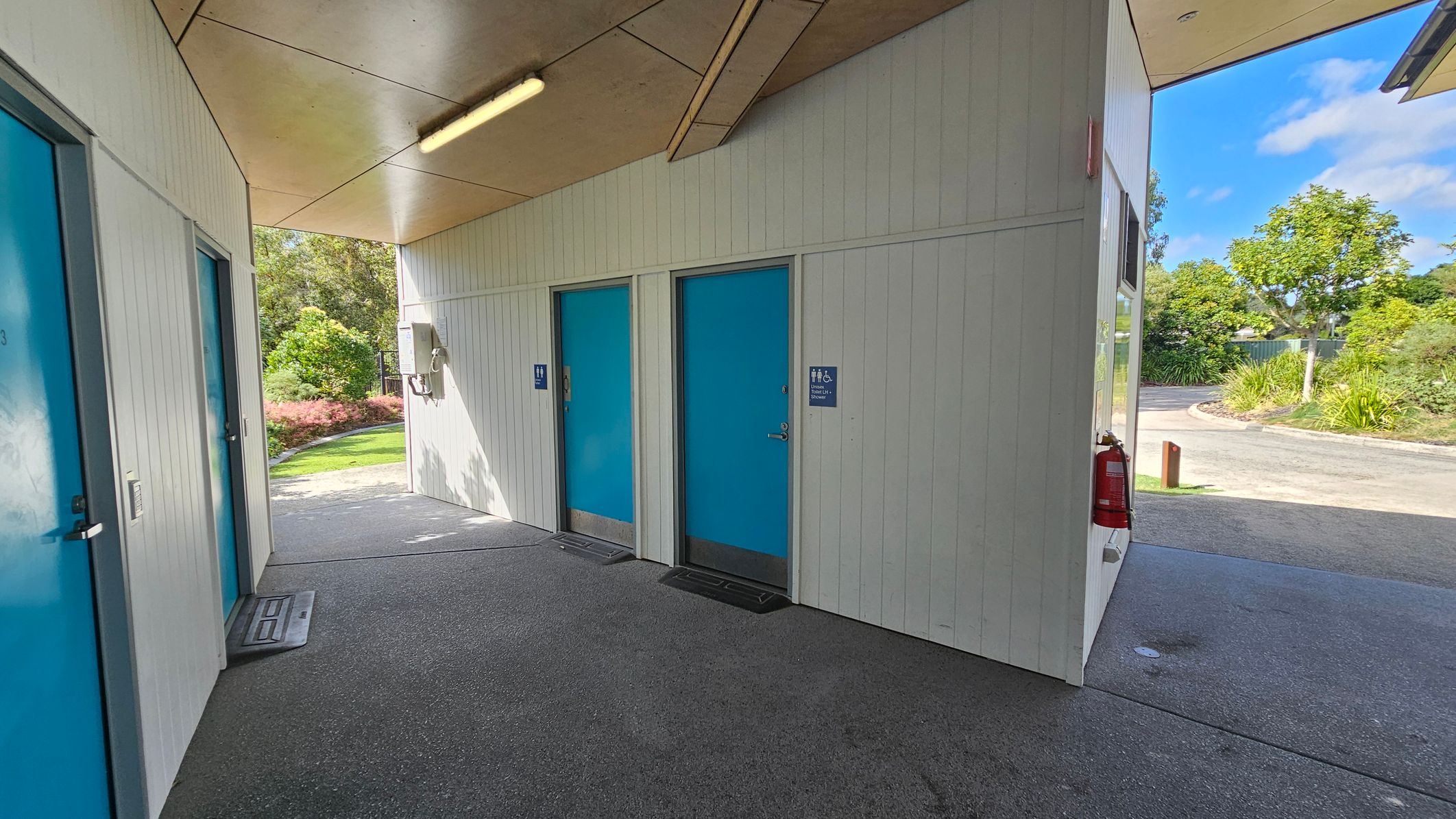The space features a grey concrete floor that extends throughout the corridor. On the left, there are two doors, one marked with a blue sign indicating male facilities, while the other has a simple blue door. A light fixture mounted on the wooden panel ceiling illuminates the area evenly. To the right, the wall continues, and a fire extinguisher is visible near the corner. Beyond the exit, greenery provides a backdrop, with shrubs and a pathway visible through an opening leading outside. The space features a grey concrete floor that extends throughout the corridor. On the left, there are two doors, one marked with a blue sign indicating male facilities, while the other has a simple blue door. A light fixture mounted on the wooden panel ceiling illuminates the area evenly. To the right, the wall continues, and a fire extinguisher is visible near the corner. Beyond the exit, greenery provides a backdrop, with shrubs and a pathway visible through an opening leading outside.