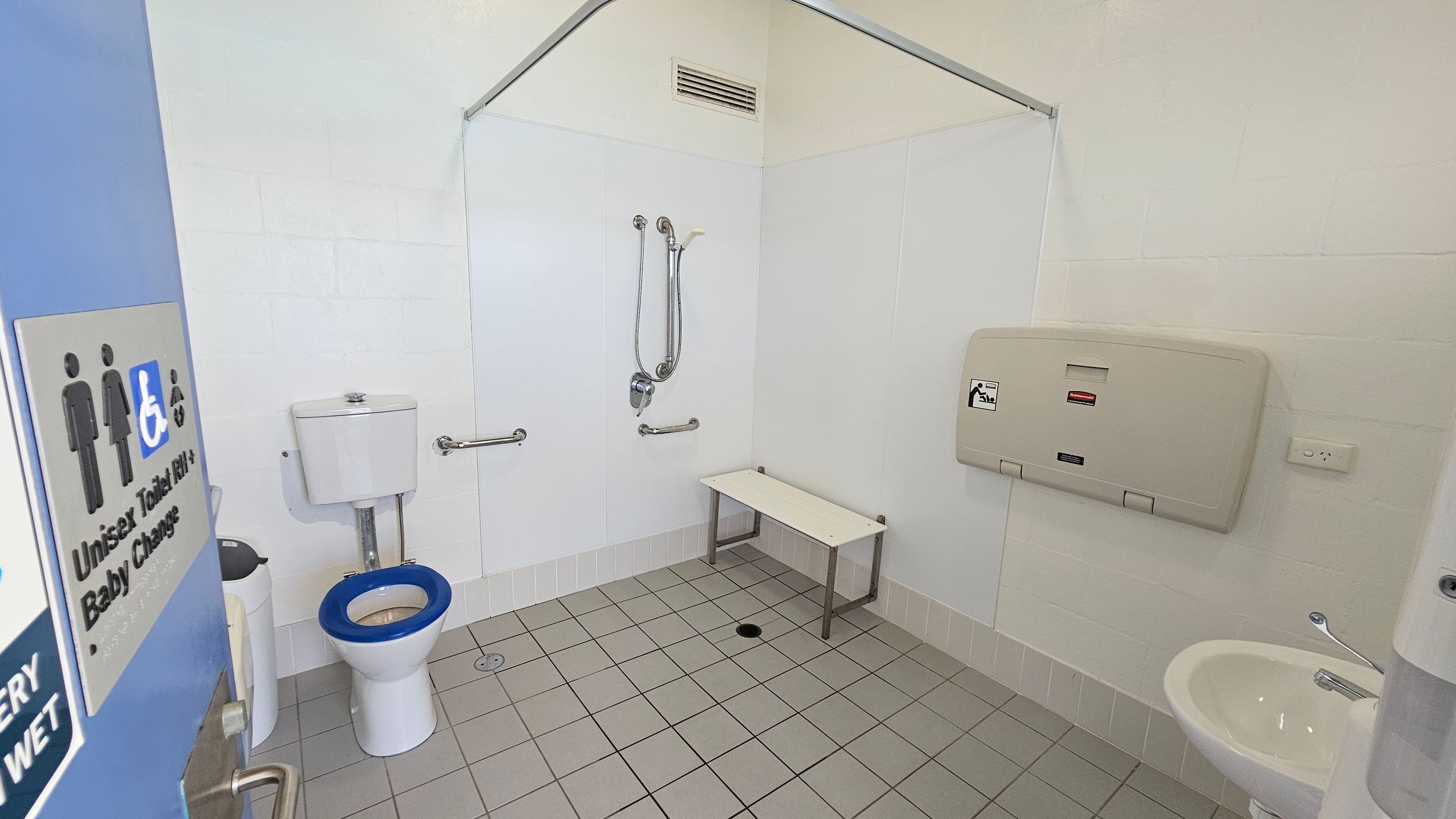 Accessible bathroom with a blue door on the left, featuring a sign indicating unisex and baby change facilities. Inside, a toilet with a blue seat is on the left, equipped with grab bars. A shower area with a handheld showerhead and additional grab bars is in the centre. A white bench is next to the shower. On the right, a baby changing station is mounted on the wall above a small sink. The floor is tiled in grey, and the walls are light-coloured. Accessible bathroom with a blue door on the left, featuring a sign indicating unisex and baby change facilities. Inside, a toilet with a blue seat is on the left, equipped with grab bars. A shower area with a handheld showerhead and additional grab bars is in the centre. A white bench is next to the shower. On the right, a baby changing station is mounted on the wall above a small sink. The floor is tiled in grey, and the walls are light-coloured.