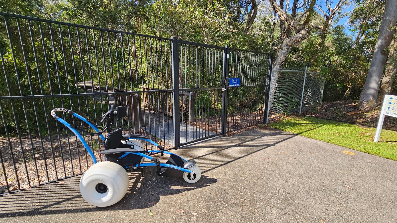 A smooth dark grey pathway leads up to a double black gate, which is partially open, allowing access to a wooded area beyond. On the left, a wheeled device sits on the path, featuring a white wheel and a blue frame, positioned close to the gate. To the right, a chain-link fence borders the pathway, and a sign indicating directions stands nearby. Lush green foliage surrounds the gate, providing a natural backdrop, while a variety of trees are visible beyond the fence. A smooth dark grey pathway leads up to a double black gate, which is partially open, allowing access to a wooded area beyond. On the left, a wheeled device sits on the path, featuring a white wheel and a blue frame, positioned close to the gate. To the right, a chain-link fence borders the pathway, and a sign indicating directions stands nearby. Lush green foliage surrounds the gate, providing a natural backdrop, while a variety of trees are visible beyond the fence.