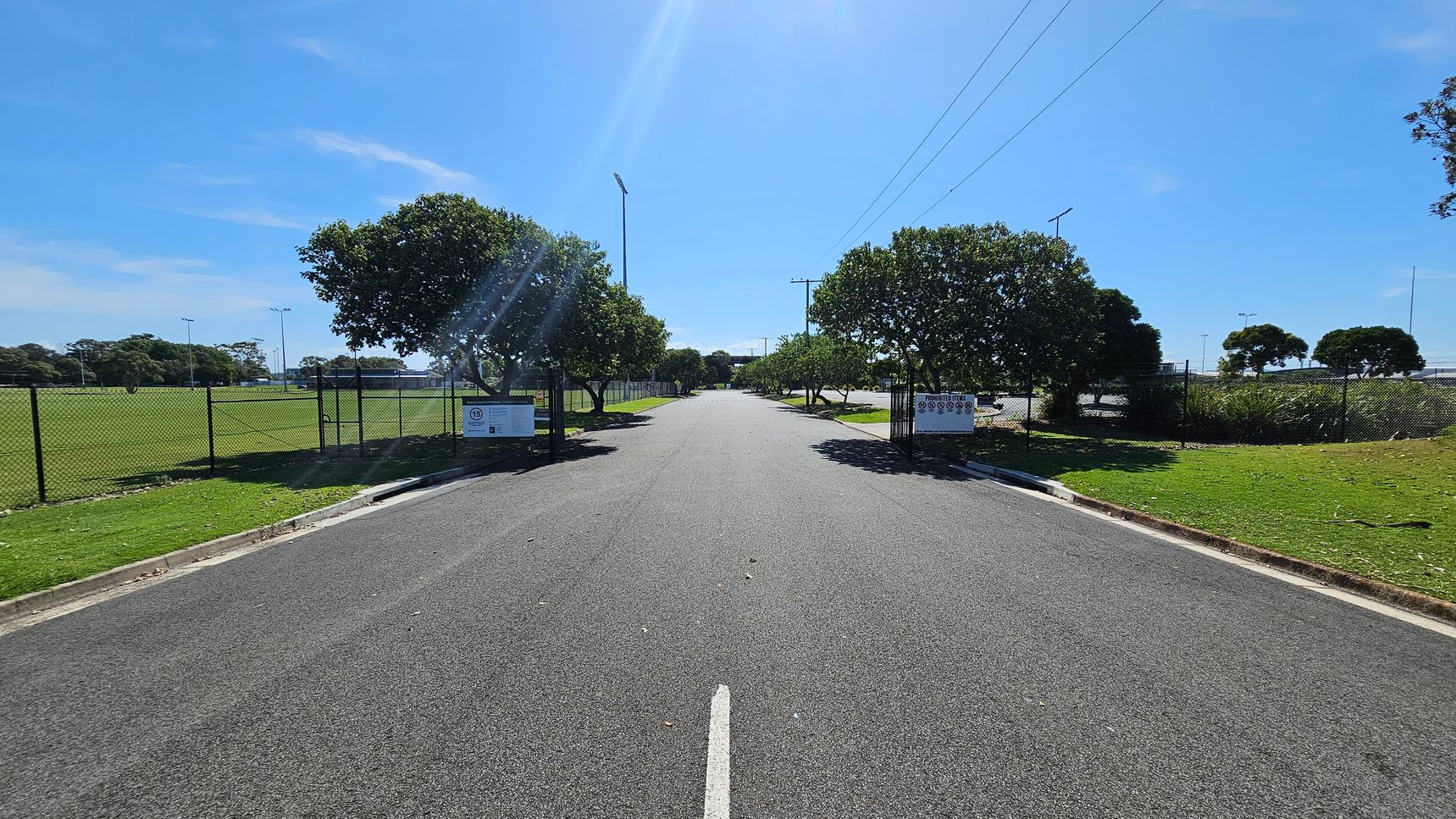 A straight, paved road flanked by green grass and trees on both sides. On the left, a chain-link fence borders a grassy field with tall light poles. On the right, another fence runs parallel to the road, with more trees and a grassy area. Signs are posted on both sides near the entrance. The sky is clear and blue, and the road has a single white line running down the centre.