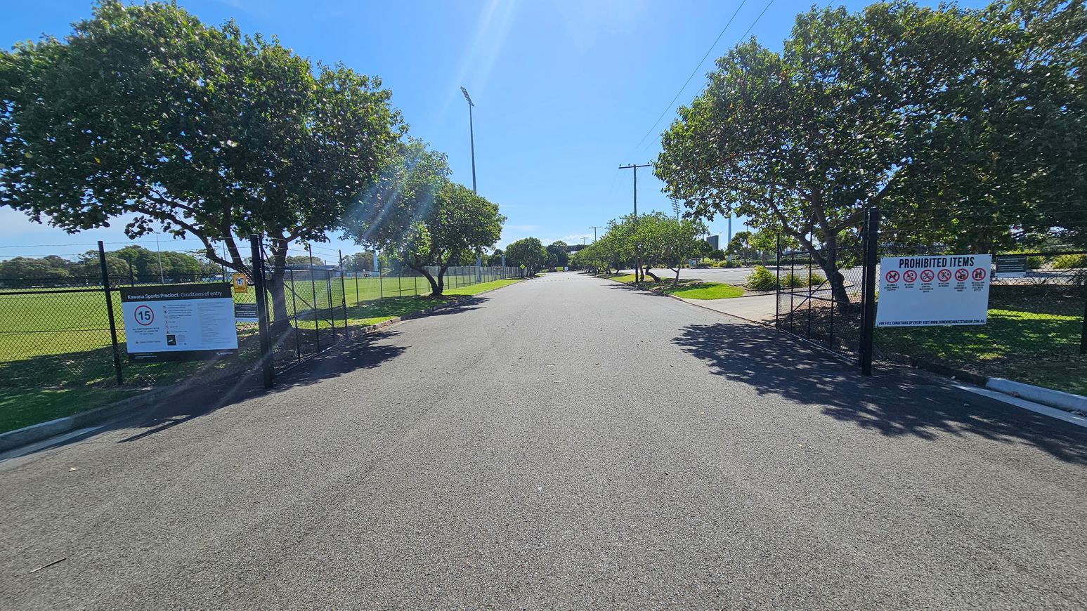 A paved road flanked by trees on both sides, leading into the distance. On the left, a chain-link fence with a sign about entry conditions. On the right, another fence with a sign listing prohibited items. The area is well-lit, with shadows from the trees cast on the ground. The road is bordered by grassy areas, and there are light poles visible in the background.