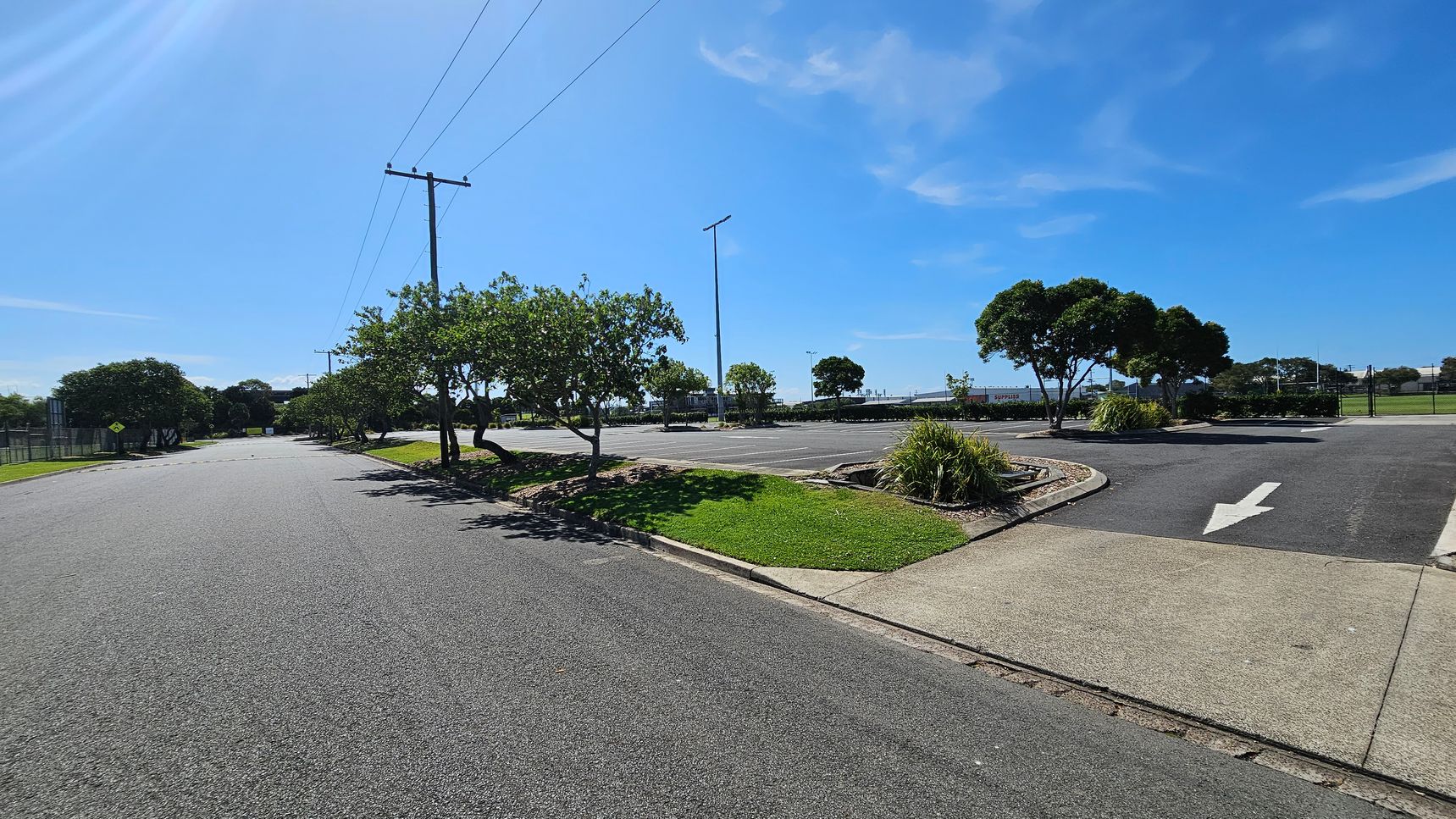 A paved road on the left curves into the distance, lined with small trees and a fence. On the right, a parking area with marked spaces and an arrow on the ground is bordered by grass and shrubs. Power lines run overhead, supported by poles. In the background, more trees and a field are visible.