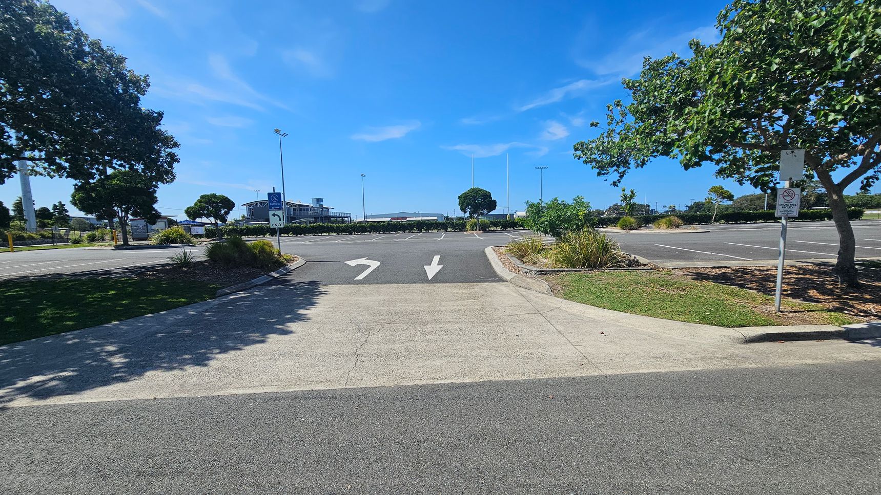 A car park with marked lanes and directional arrows on the asphalt. Trees line both sides, providing shade. A signpost on the right indicates parking regulations. In the background, a building is visible, along with more trees and light poles. The ground is a mix of asphalt and concrete, with patches of grass and small shrubs along the edges.