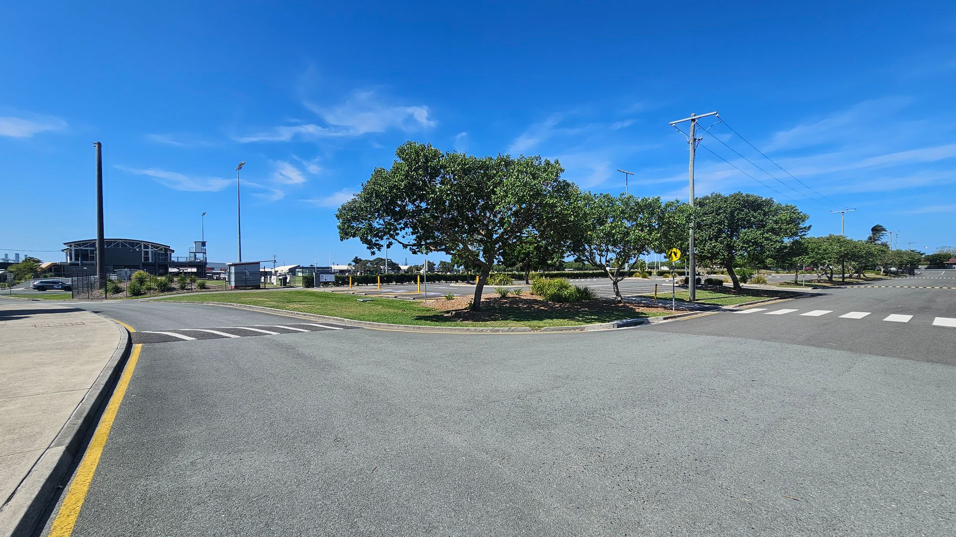 A wide road intersection with a pedestrian crossing on the right. In the centre, there is a grassy median with several trees. On the left, a building with a curved roof is visible, along with a fenced area and a parked car. Power lines run along the right side of the road. The pavement is grey, and the kerb has a yellow line.