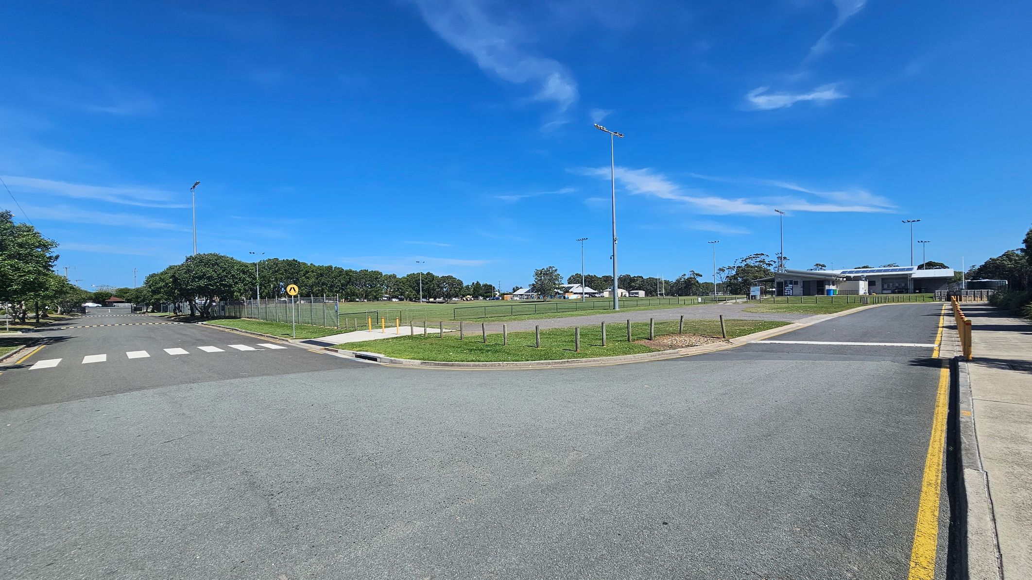 A paved road with a zebra crossing on the left leads to a grassy area with a fenced sports field. Tall floodlights are visible around the field. On the right, a modern building with large windows is situated near the road, surrounded by more grass and trees. The road curves slightly, bordered by a yellow line and a footpath. Trees line the background, and the sky is clear.