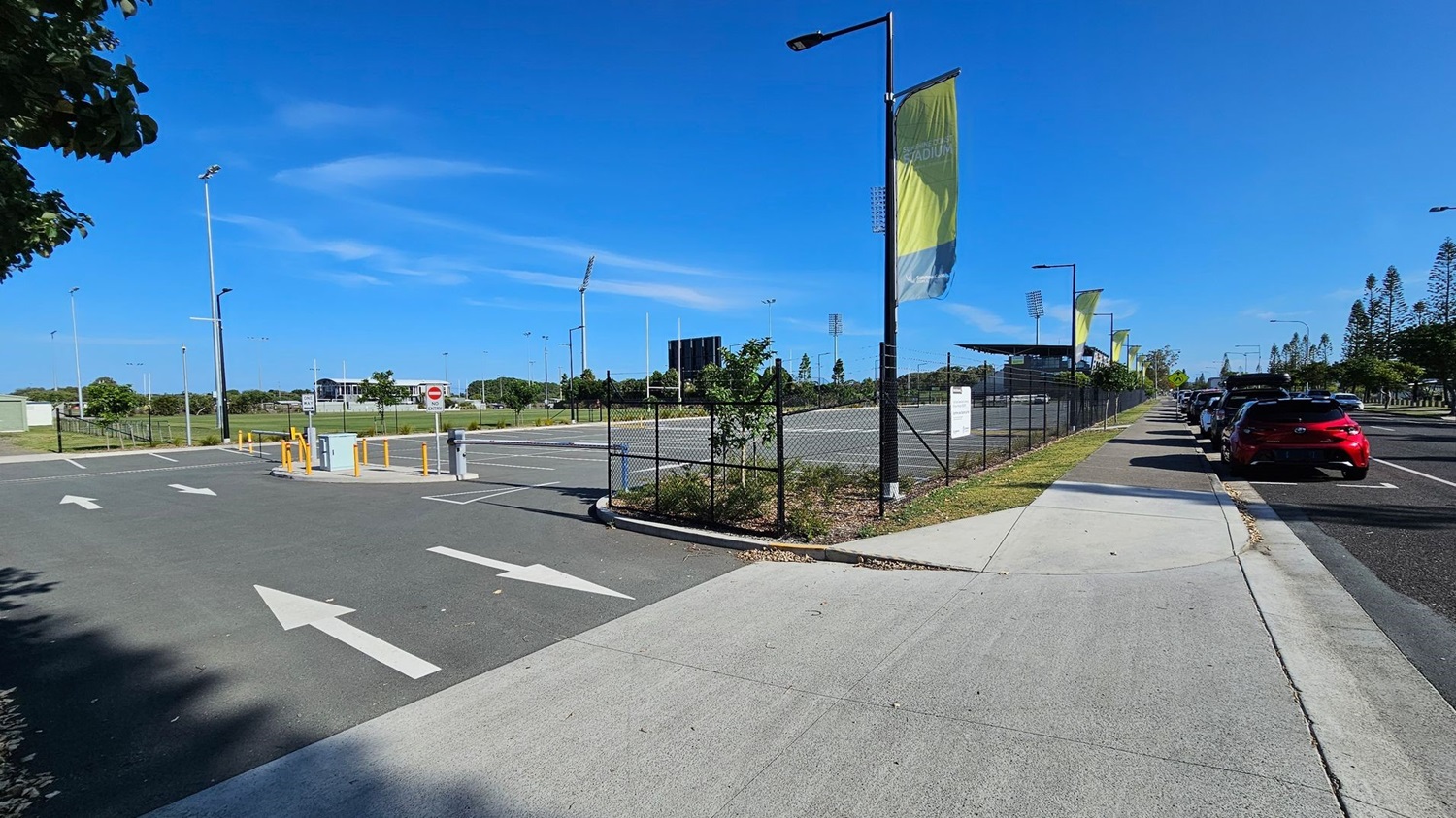 A paved road with white directional arrows is in the foreground, leading to a fenced area on the left. Tall floodlights and a scoreboard are visible in the background, suggesting a sports field. On the right, a row of parked cars lines the street, with a red car closest to the foreground. Banners hang from streetlights along the pavement, and trees are visible in the distance. The sky is clear and blue.