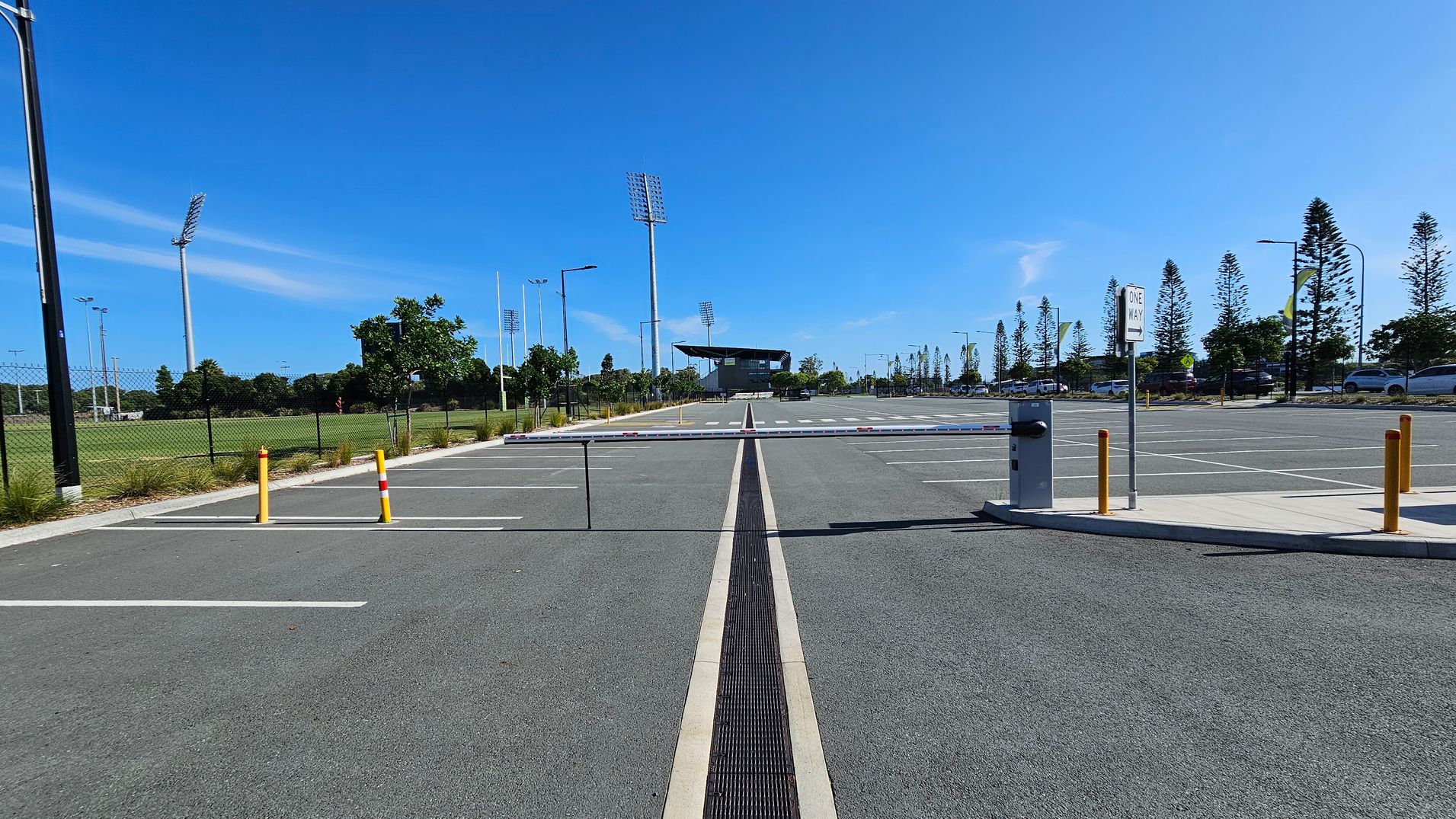 A large, empty car park with marked spaces and a central barrier gate. On the left, there's a grassy sports field with tall floodlights and a chain-link fence. On the right, a "One Way" sign and several parked cars are visible. In the background, a structure resembling a stadium or sports facility is surrounded by trees and more floodlights. Tall, narrow trees line the horizon. The ground is paved with asphalt.