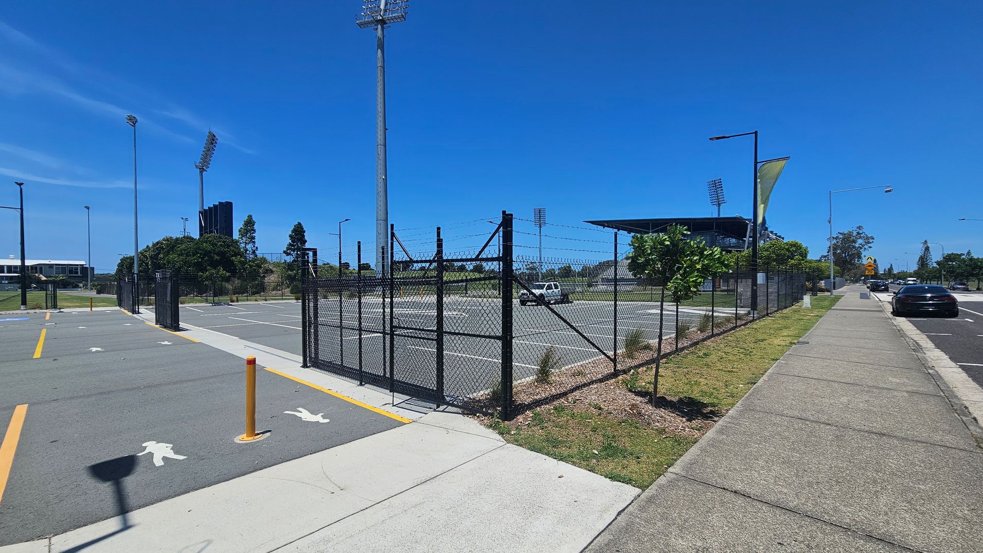 A paved walkway runs along the right side, bordered by a grassy strip with small trees. A black chain-link fence with barbed wire encloses a parking area on the left, featuring painted pedestrian symbols and yellow lines. Tall floodlights and a stadium structure are visible in the background. A few cars are parked along the street on the right. The scene is well-lit, suggesting a sunny day.