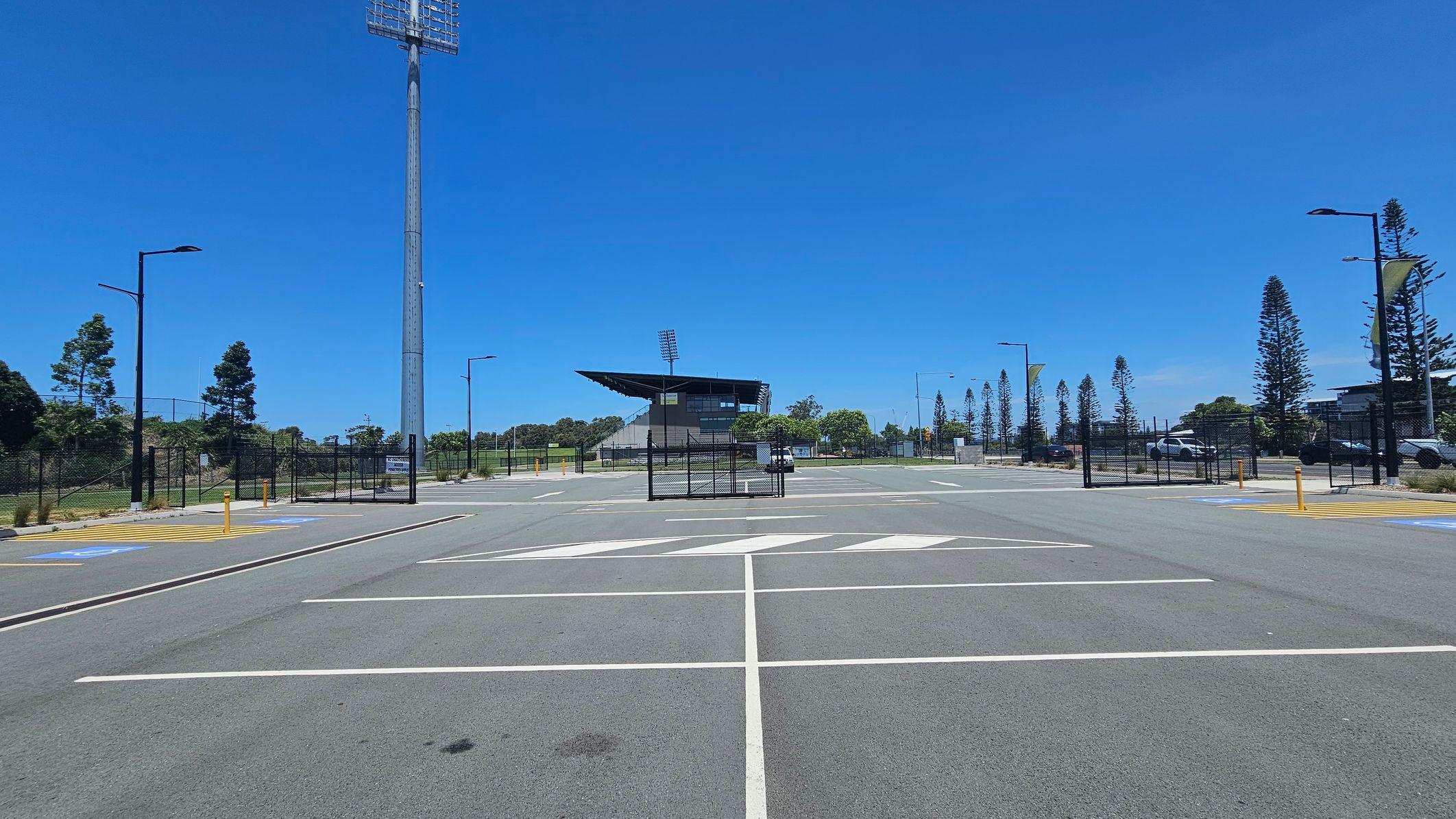 An empty car park with marked spaces and a large arrow on the asphalt. In the centre, a tall floodlight tower stands near a fenced area. Beyond the fence, a stadium with a distinctive roof is visible. Trees and lampposts line the sides, with a few cars parked on the right. The sky is clear and blue.
