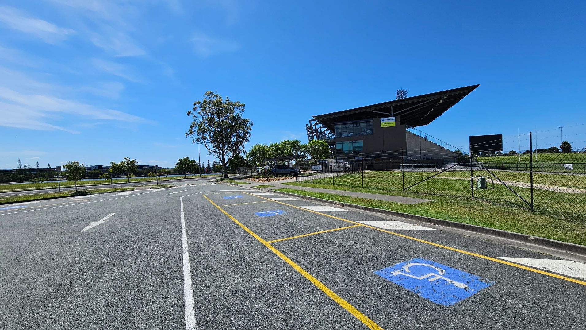 A large sports stadium is on the right, with a modern design and a prominent overhanging roof. In the foreground, there is a car park with marked disabled parking spaces, indicated by blue symbols on the asphalt. A chain-link fence runs along the perimeter of the stadium grounds. Trees and grass are visible around the area, with a few small trees lining the road on the left. The road has directional arrows painted on it.