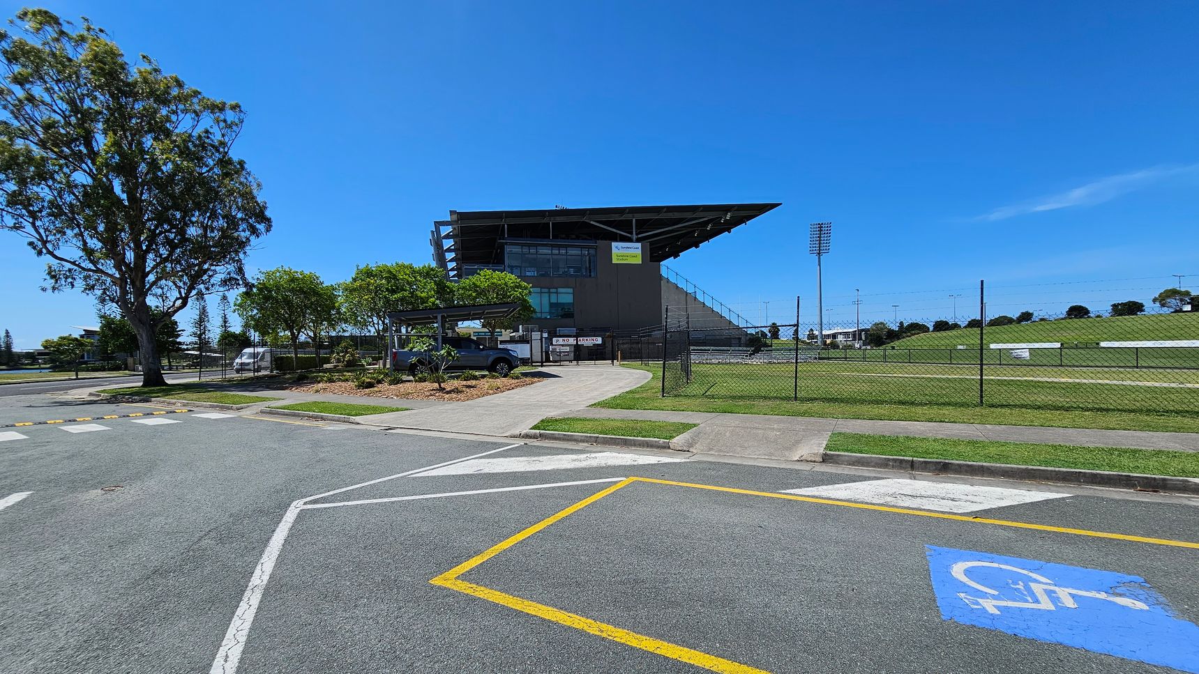A sports stadium with a large, angular roof is in the centre. To the left, there is a tall tree and a few smaller trees near a parked car. A fence runs along the right side, enclosing a grassy field with floodlights. The foreground shows a car park with a marked disabled parking space and a pedestrian crossing. The sky is clear and blue.
