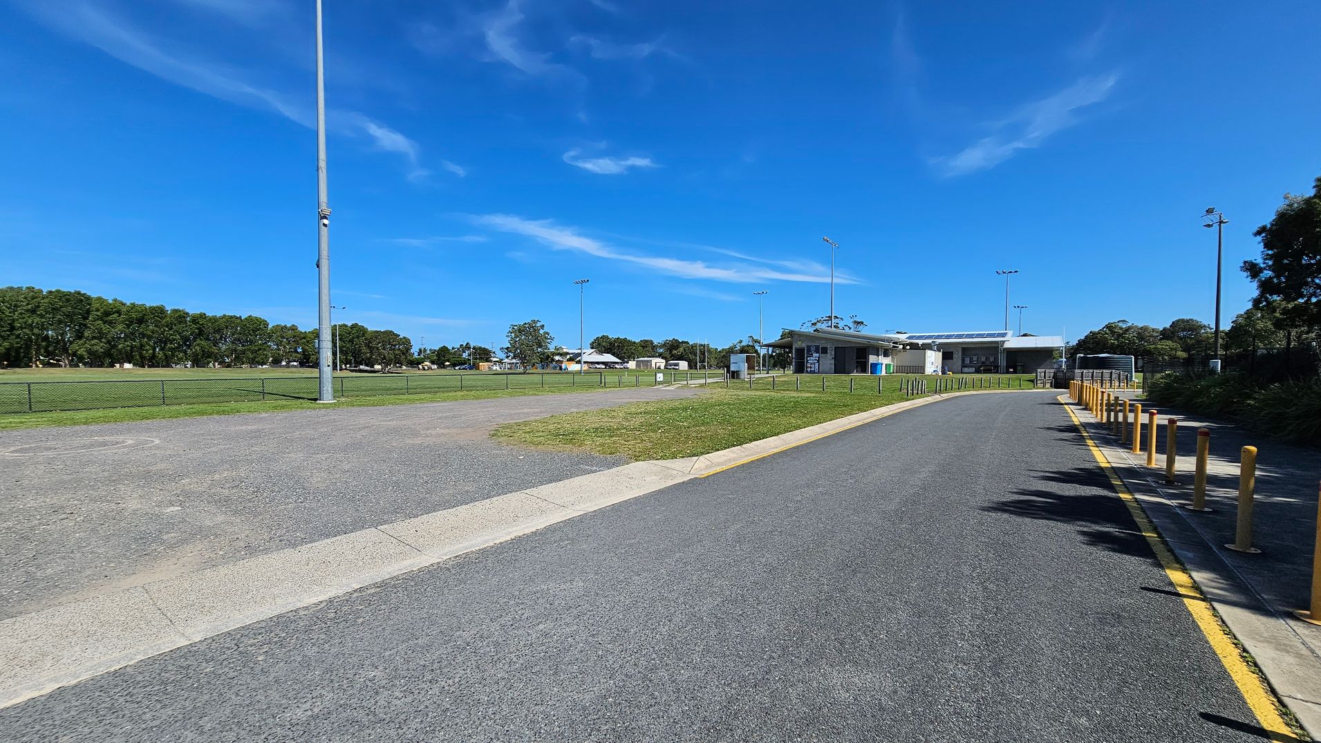 A paved road on the right with yellow bollards leads to a building in the distance. On the left, there's a grassy field bordered by a low fence and a tall light pole. Trees line the background, and the sky is clear.