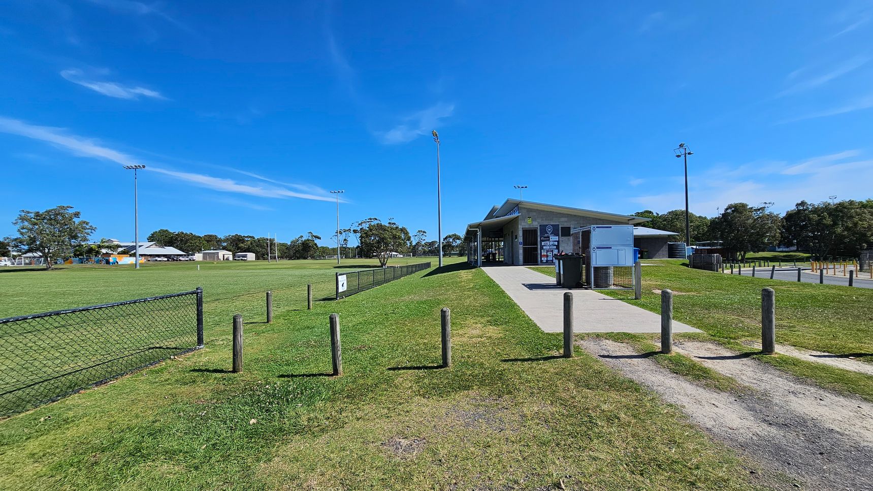 A grassy sports field with a chain-link fence on the left. In the centre, a path leads to a building with a sloped roof and several signs on its walls. Tall floodlights are visible around the field. Wooden posts line the path, and there are trees and additional buildings in the background. The sky is clear and blue.