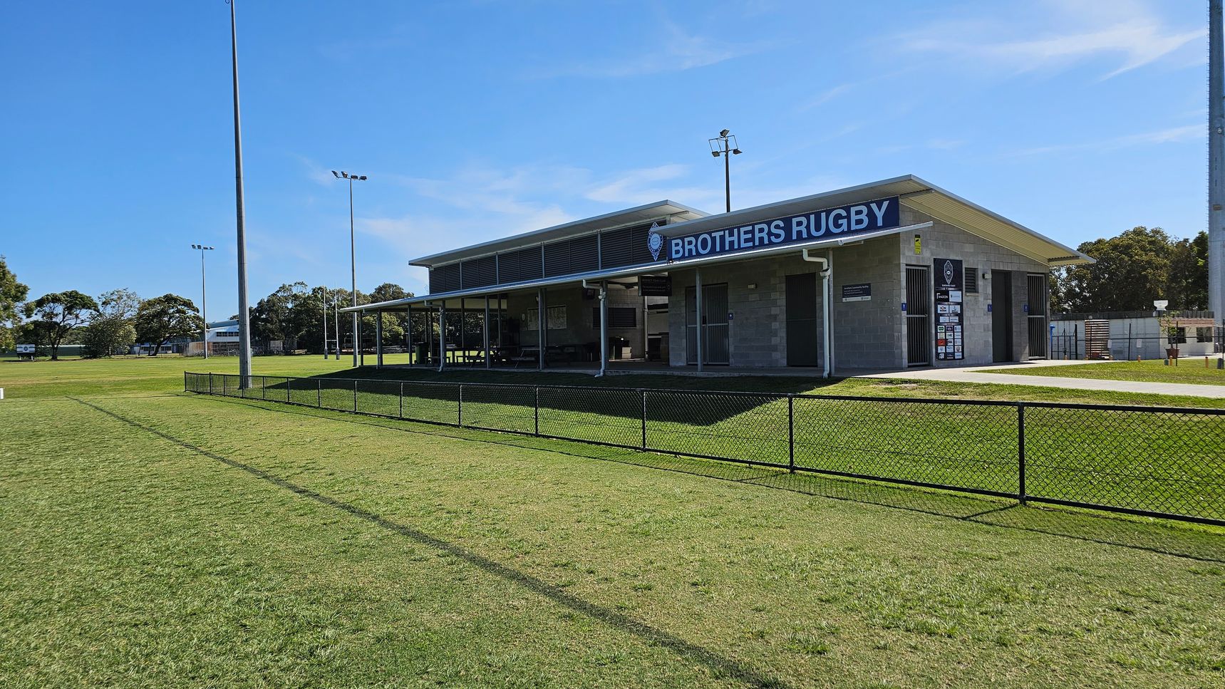 A rugby field with a clubhouse on the right, labelled "Brothers Rugby". The building is single-storey with a flat roof and has a covered area with seating. A black chain-link fence runs along the foreground, separating the field from the clubhouse. Tall floodlights are visible around the field, and trees are in the background. The grass is well-maintained, and the sky is clear.