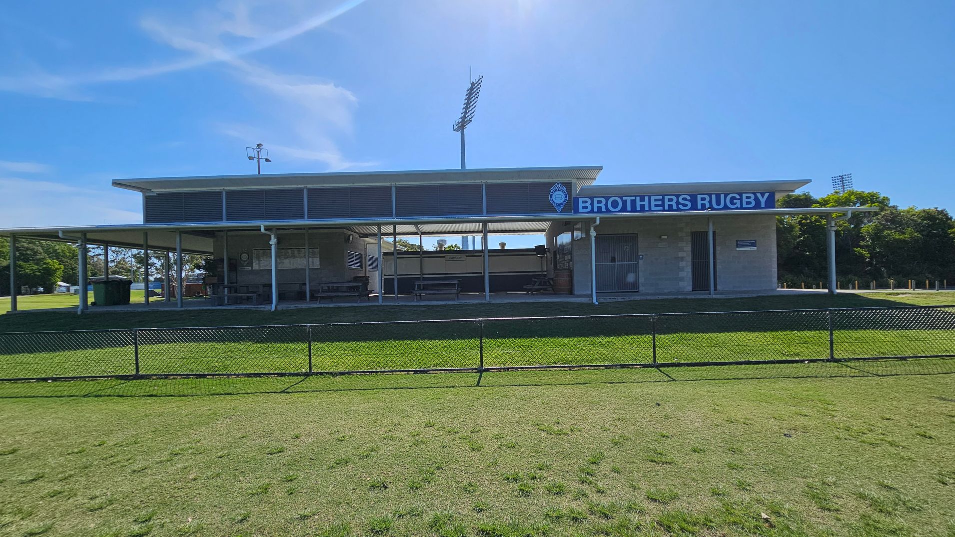A rugby clubhouse with a sign reading "Brothers Rugby" is in the centre. The building has a flat roof and open sides with benches underneath. A tall floodlight stands behind it. The foreground features a grassy field with a low chain-link fence. Trees are visible on the right and left sides in the background.