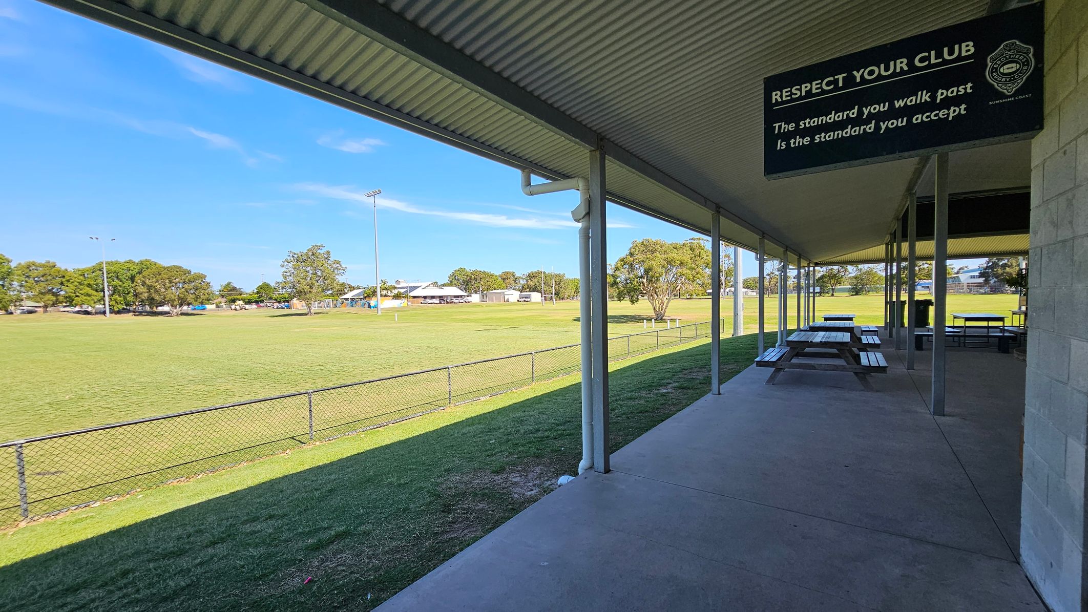A sports field with a grassy area and a chain-link fence on the left. In the middle, there are trees and a few buildings in the distance. On the right, a covered area with a corrugated metal roof, featuring picnic tables and a sign that reads, "RESPECT YOUR CLUB. The standard you walk past is the standard you accept." The ground is concrete, and the ceiling is metal.