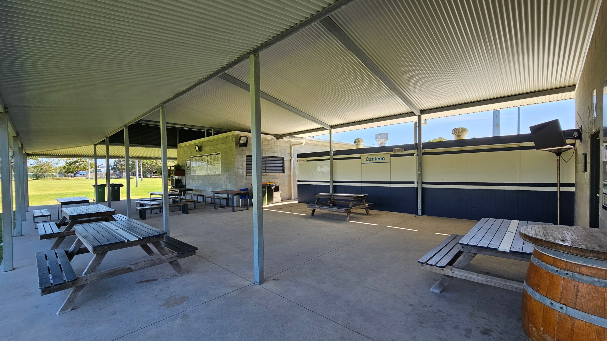A covered outdoor area with a corrugated metal roof and concrete floor. On the left, there are several wooden picnic tables and benches. In the centre, a brick wall with a noticeboard and a canteen window. On the right, a wooden barrel and more seating. The background shows a grassy field with trees.