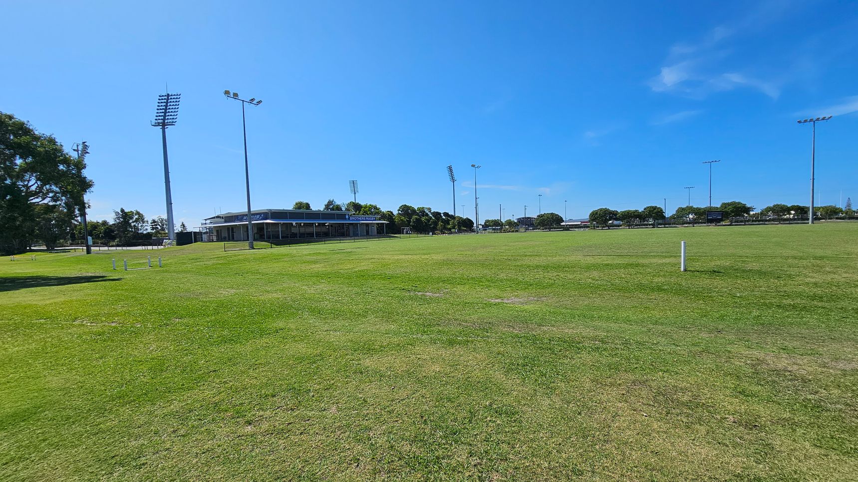 A large grassy sports field with several tall floodlights scattered around. On the left, there are trees and a building with a sign that reads "Brothers Rugby". In the background, more trees and additional floodlights are visible. The sky is clear and blue.