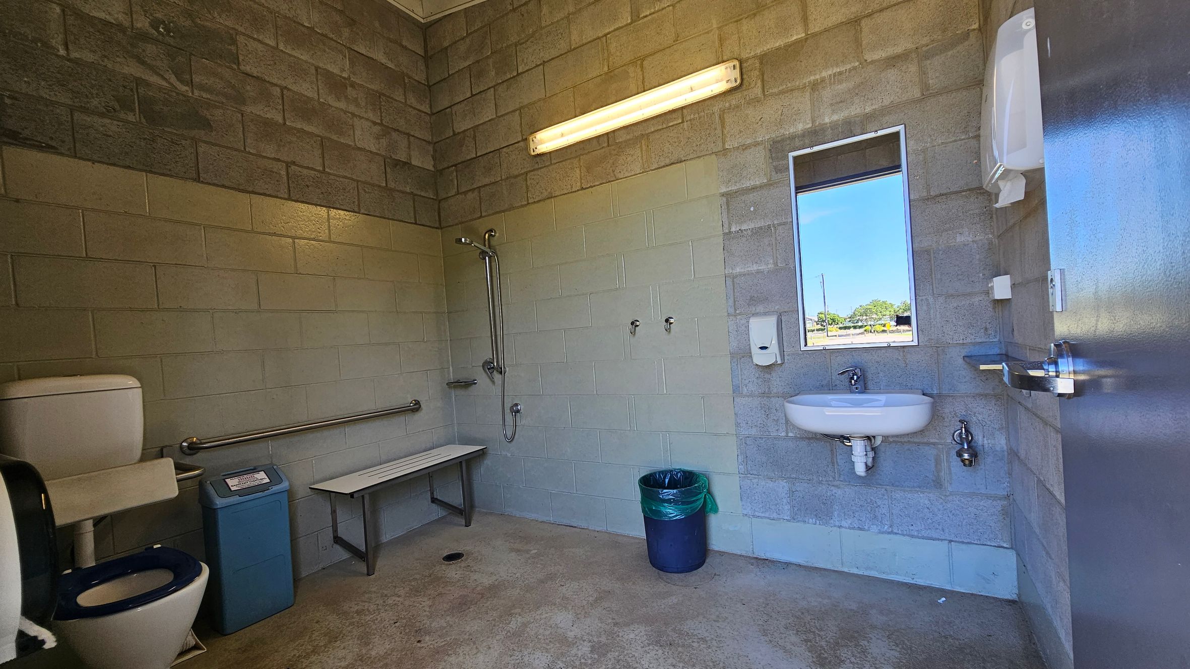 A public restroom with concrete block walls and a concrete floor. On the left, there's a toilet with a blue seat, a grab bar, and a blue bin. A shower area with a bench and additional grab bars is nearby. On the right, a sink with a mirror above it and a soap dispenser is mounted on the wall. A green bin is on the floor. A fluorescent light fixture is mounted on the wall above the shower area. A metal door is partially visible on the right.