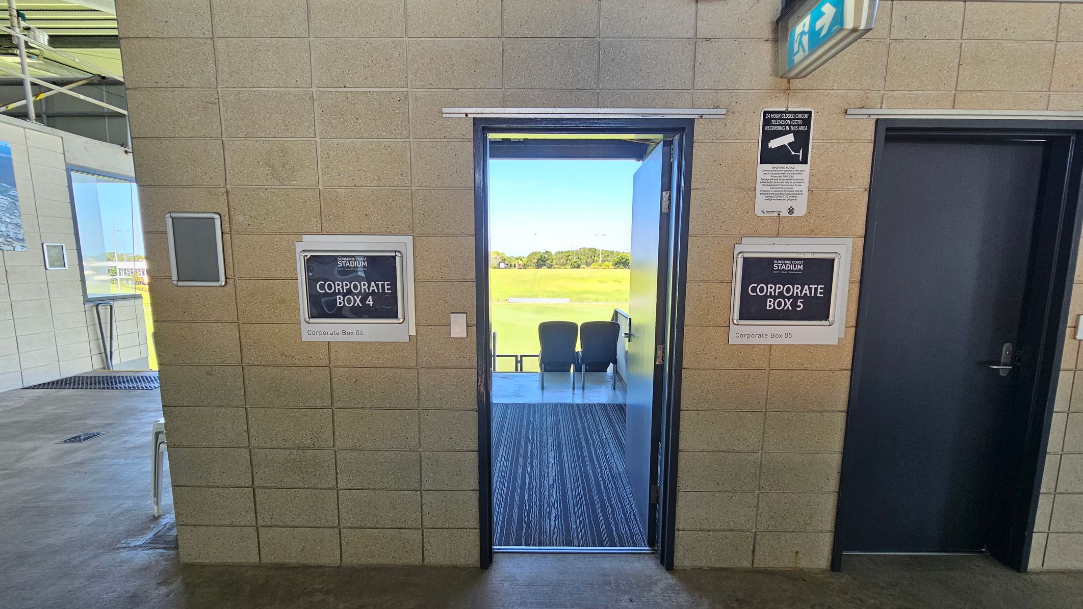 A corridor with beige tiled walls. In the centre, an open door leads to Corporate Box 4, revealing two chairs facing a sports field. To the right, a closed door marked Corporate Box 5. Above, a CCTV sign and an emergency exit sign. On the left, part of another room with a window and a chair visible. The floor is concrete, and the ceiling has visible metal beams.