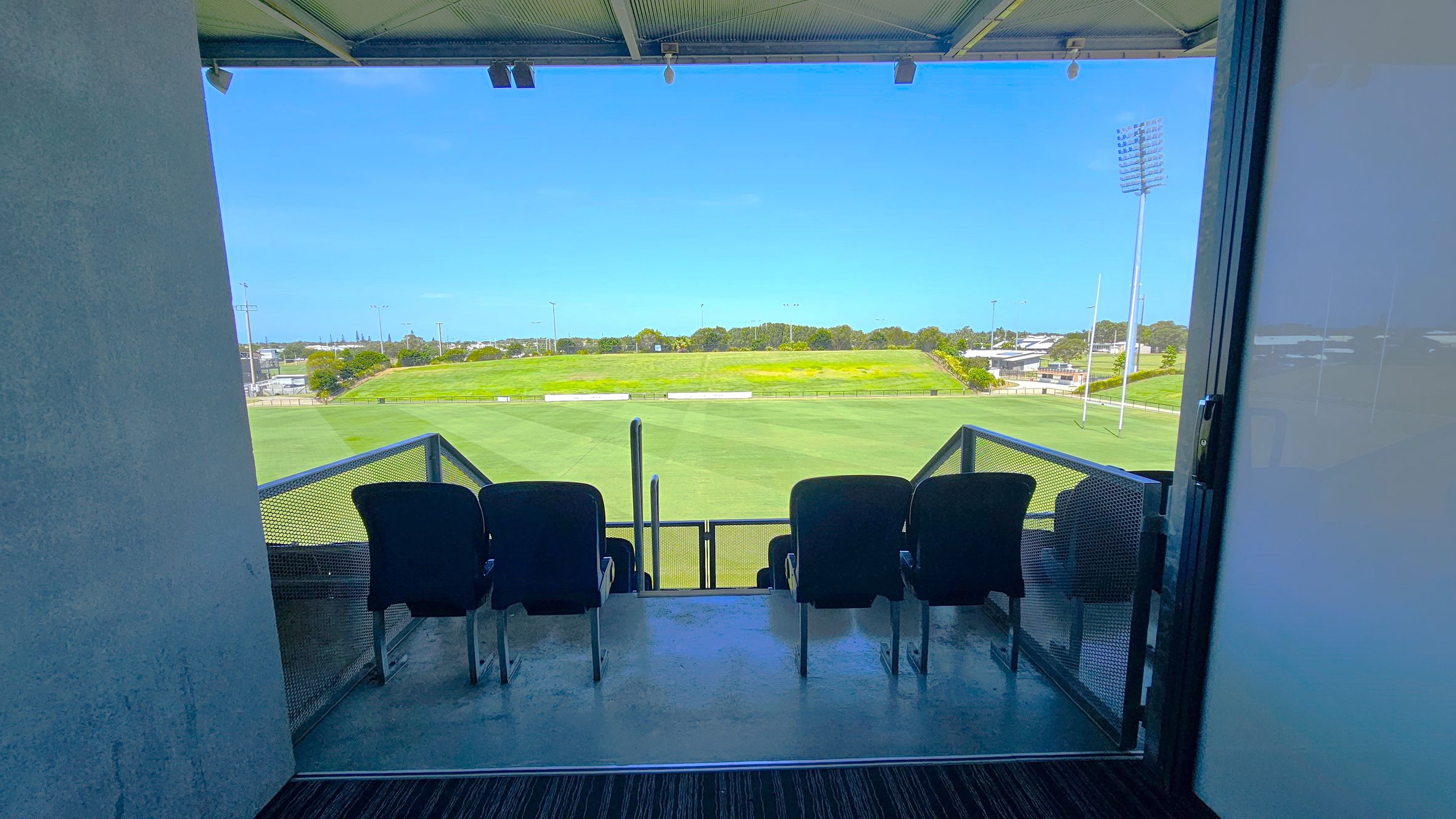 A view from inside a room looking out onto a sports field. In the foreground, there are four black seats on a small balcony with metal railings. The field is expansive and green, with goalposts visible on the right. In the background, there are trees and some buildings. The ceiling inside the room is metallic, and the floor is carpeted.