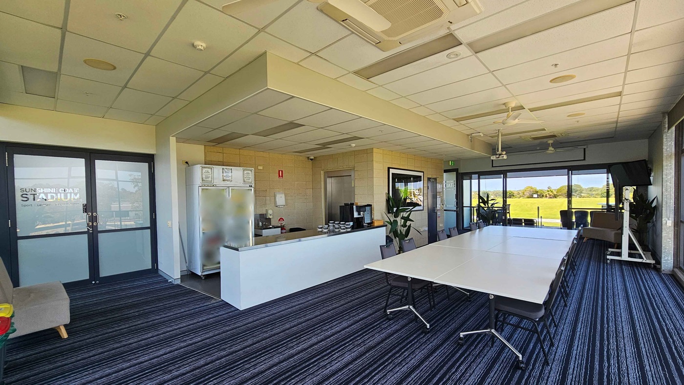 A room with a striped carpet and a grid-patterned ceiling. On the left, there are double doors with "Sunshine Coast Stadium" written on them. A grey sofa is nearby. In the centre, a counter with a coffee machine and a fridge stocked with drinks. On the right, a long table with chairs faces large windows showing a grassy field outside. A TV is mounted on a stand near the windows, and potted plants are placed around the room.