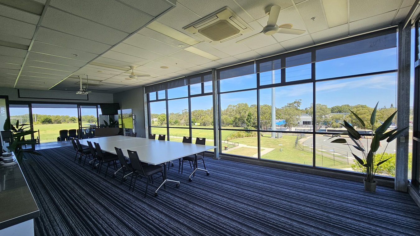 A conference room with large floor-to-ceiling windows on the right, offering a view of a green landscape and trees. In the centre, there is a long white table surrounded by black chairs on a striped carpet. On the left, there is a counter with cups and a plant. The ceiling has a projector and ceiling fans. A potted plant is near the windows.